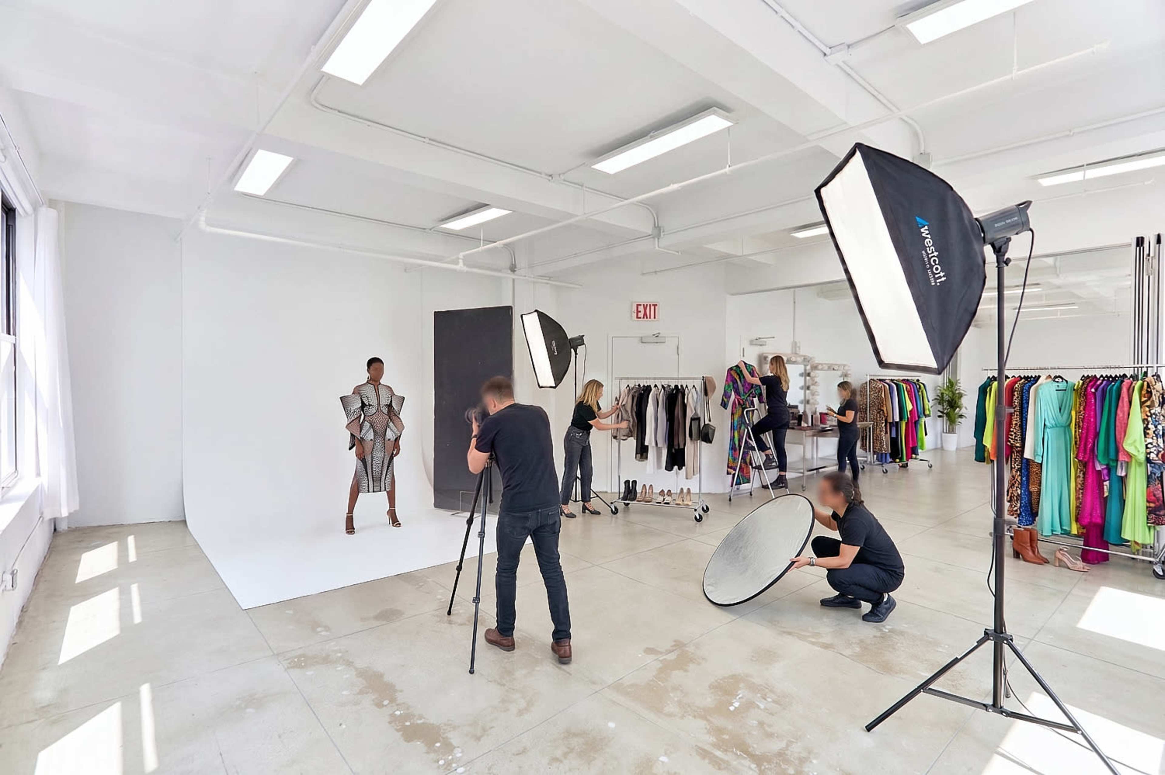 A fashion photoshoot is taking place in a studio, with a model posing in a distinctive outfit while photographers and assistants set up lighting and equipment.