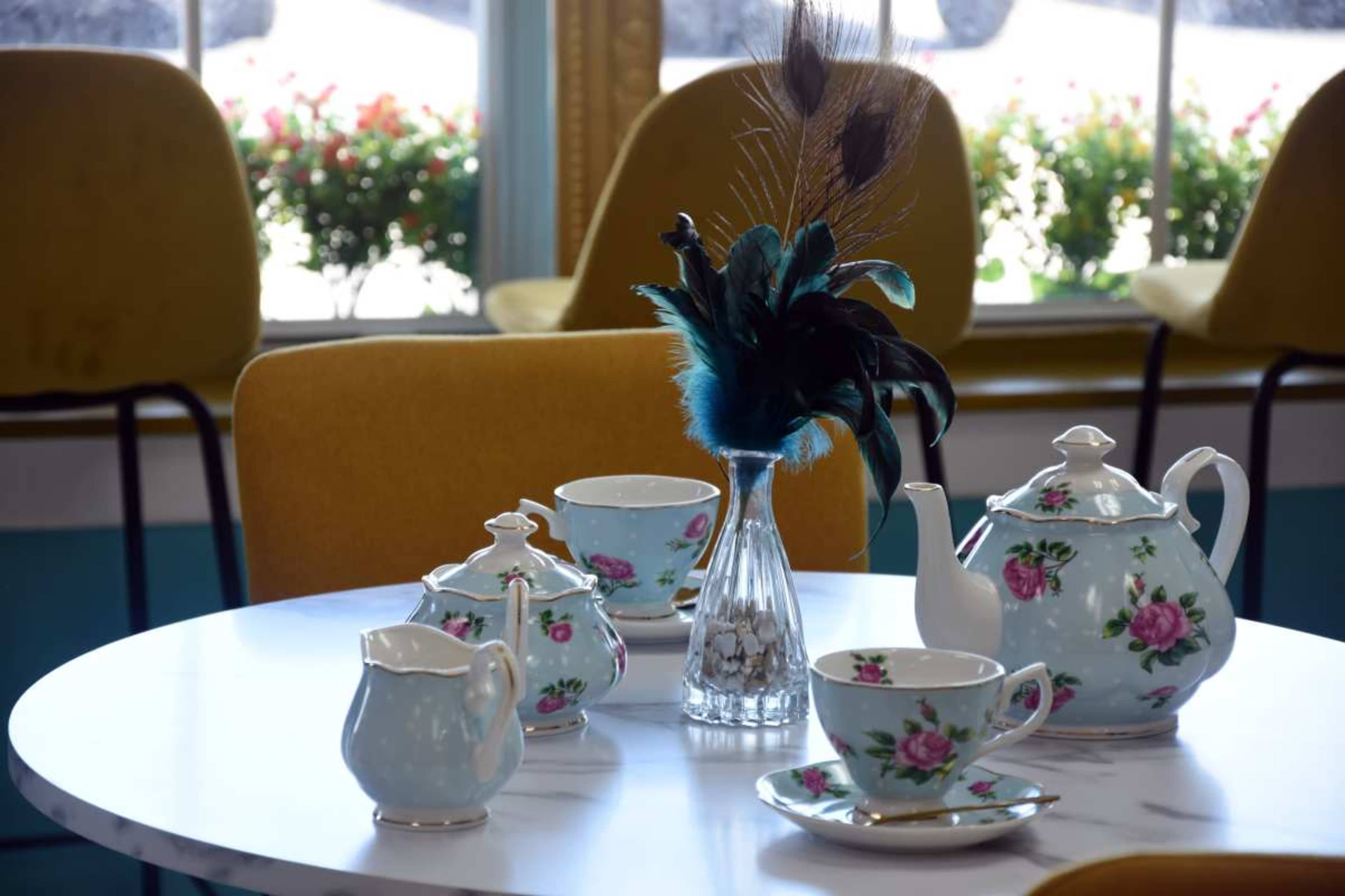 A floral-patterned tea set is arranged on a round table with a blue vase holding peacock feathers as a centerpiece.