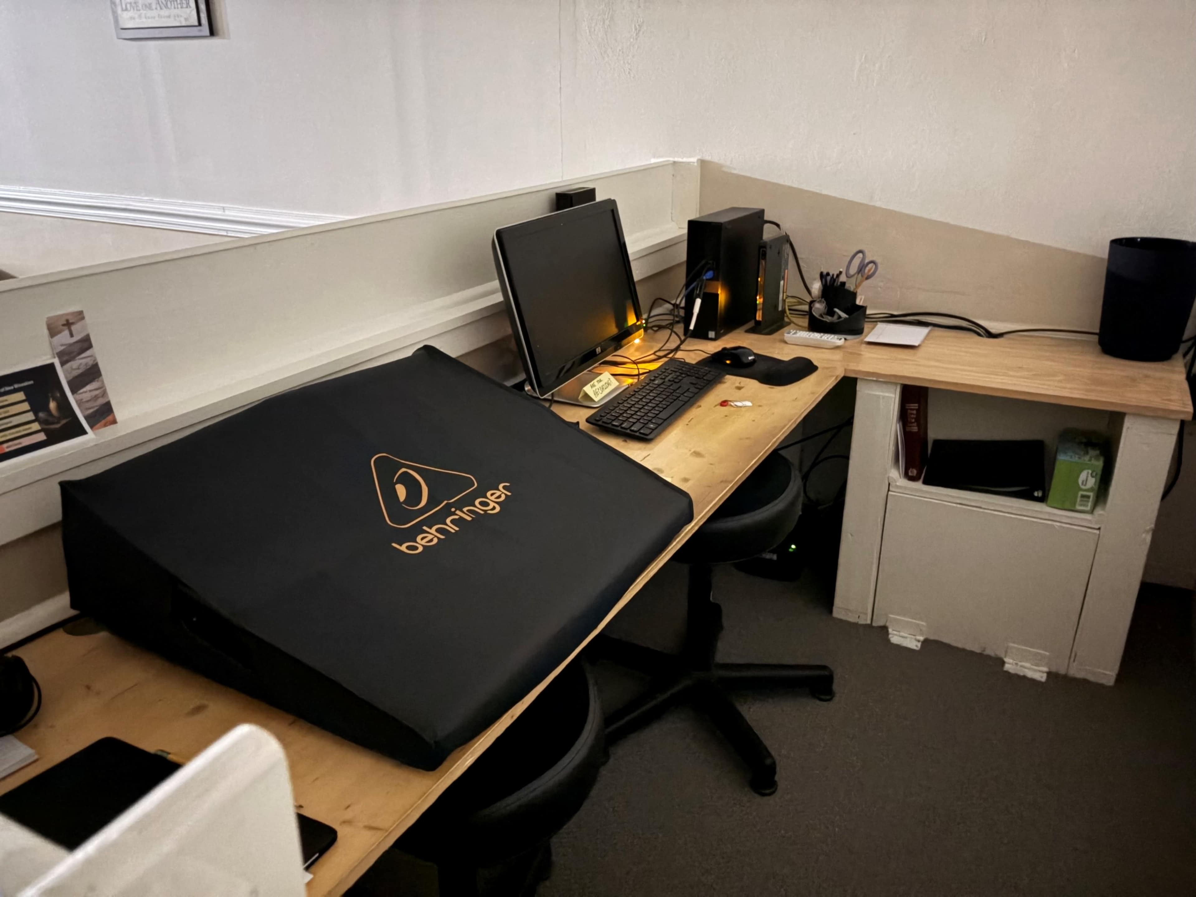 The image features an office workspace with a computer setup, including a monitor, keyboard, and speakers, alongside a large black cover displaying the Behringer logo.