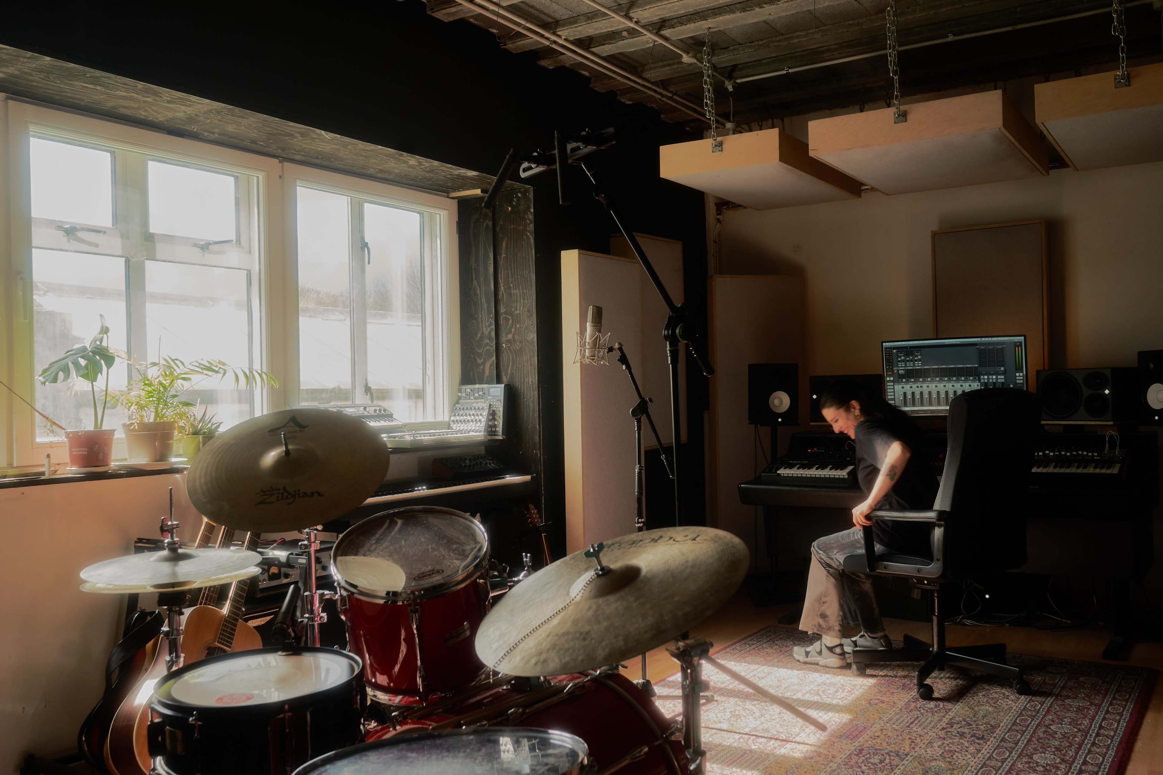 A musician sits in a chair in a recording studio, surrounded by musical instruments and equipment, with sunlight streaming through a window.