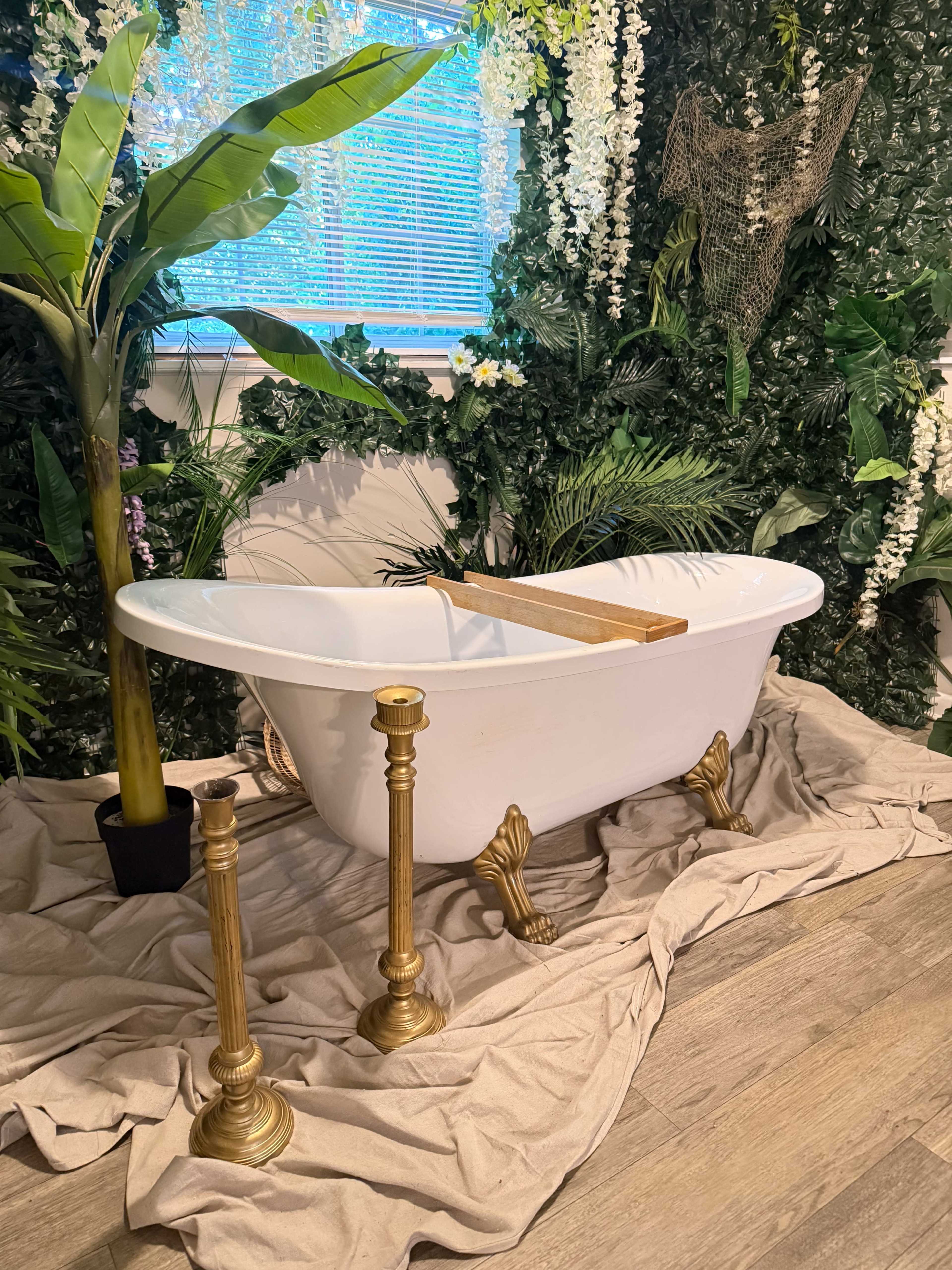 A vintage clawfoot bathtub is positioned on a draped fabric surface, surrounded by lush green plants and cascading white flowers.