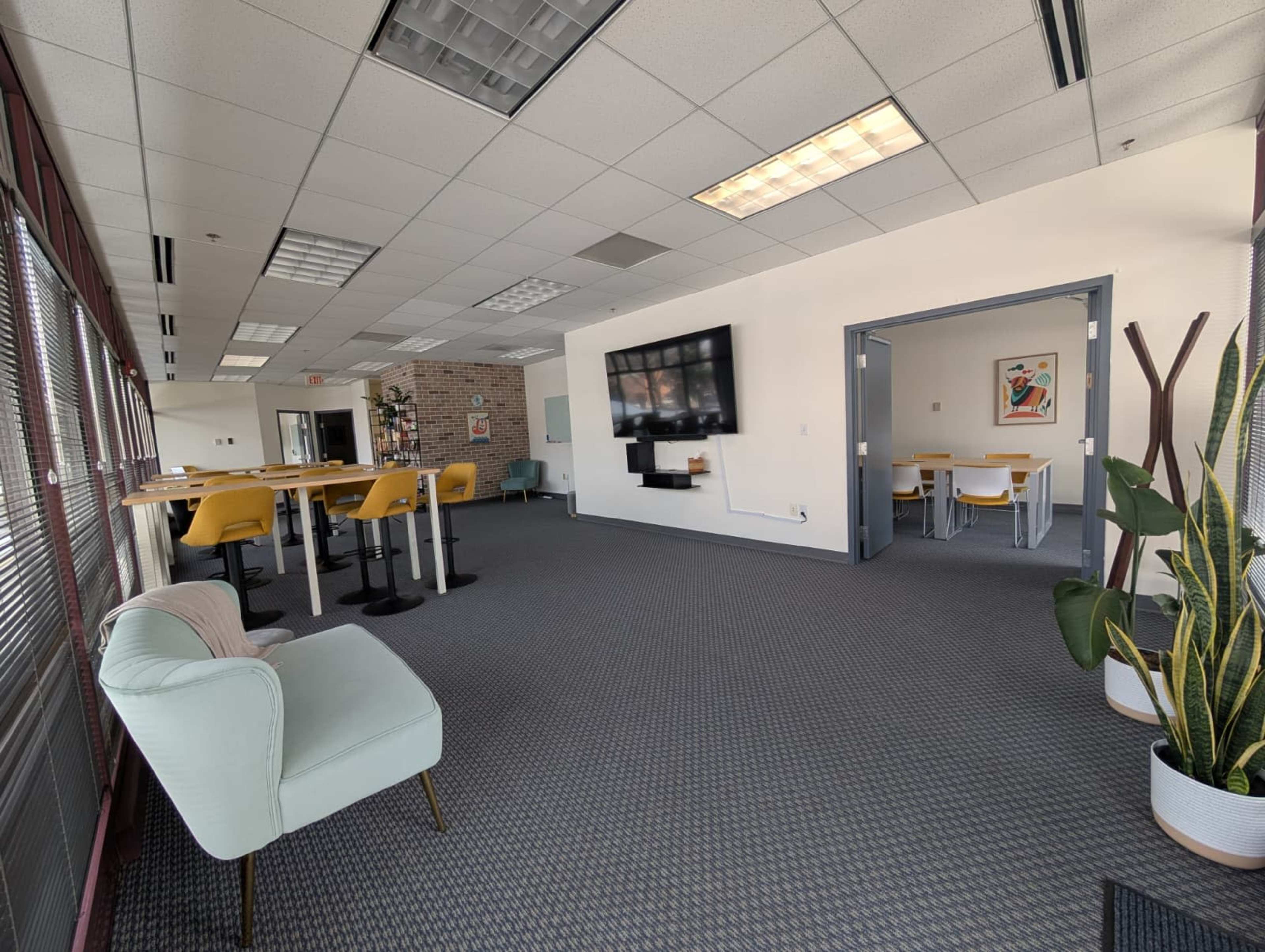 The image shows a modern office space with a seating area, high tables, and a wall-mounted television, along with plants and artwork.