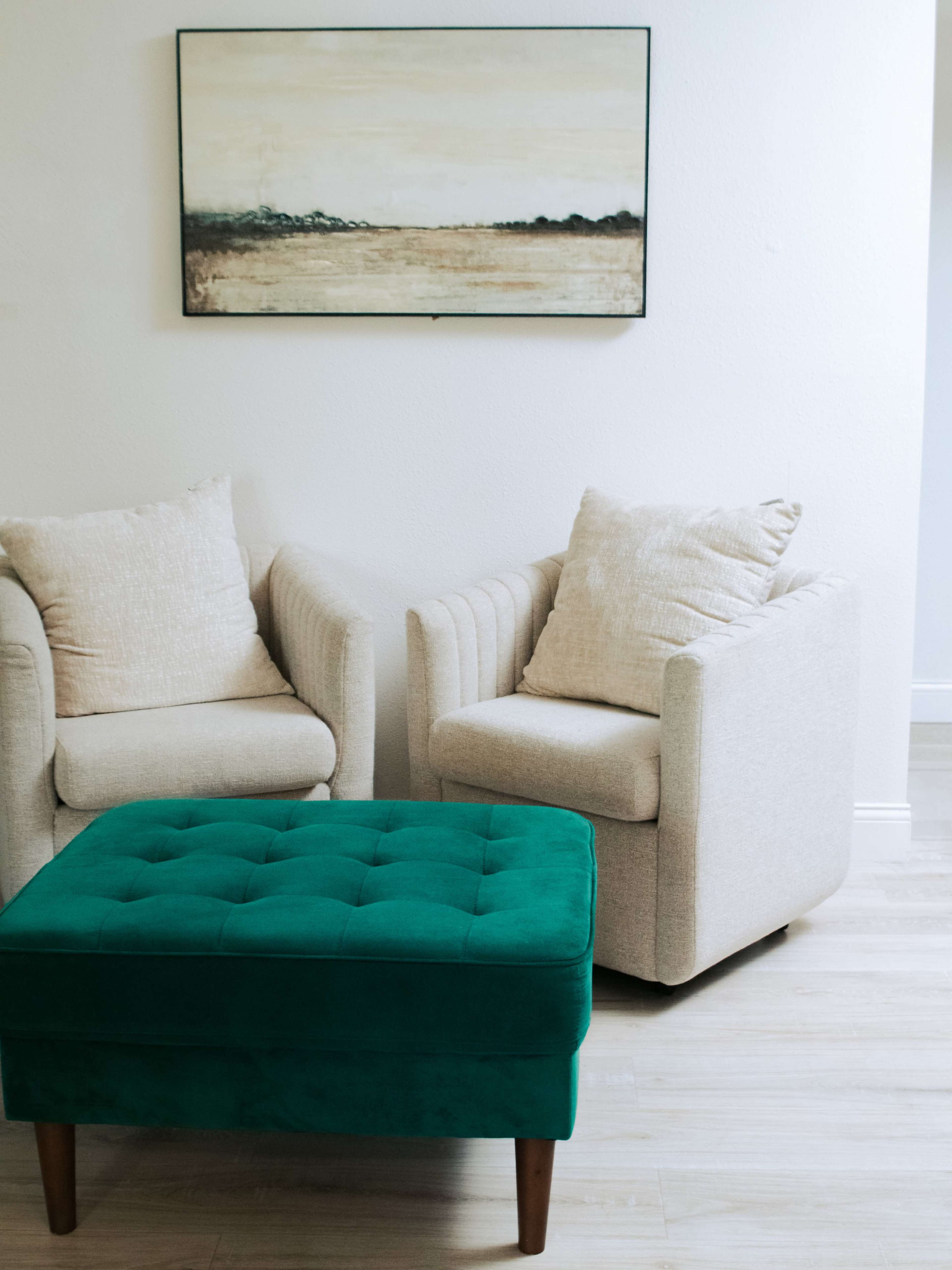 Two armchairs with cushions and a green tufted ottoman are positioned near a wall with a landscape painting.