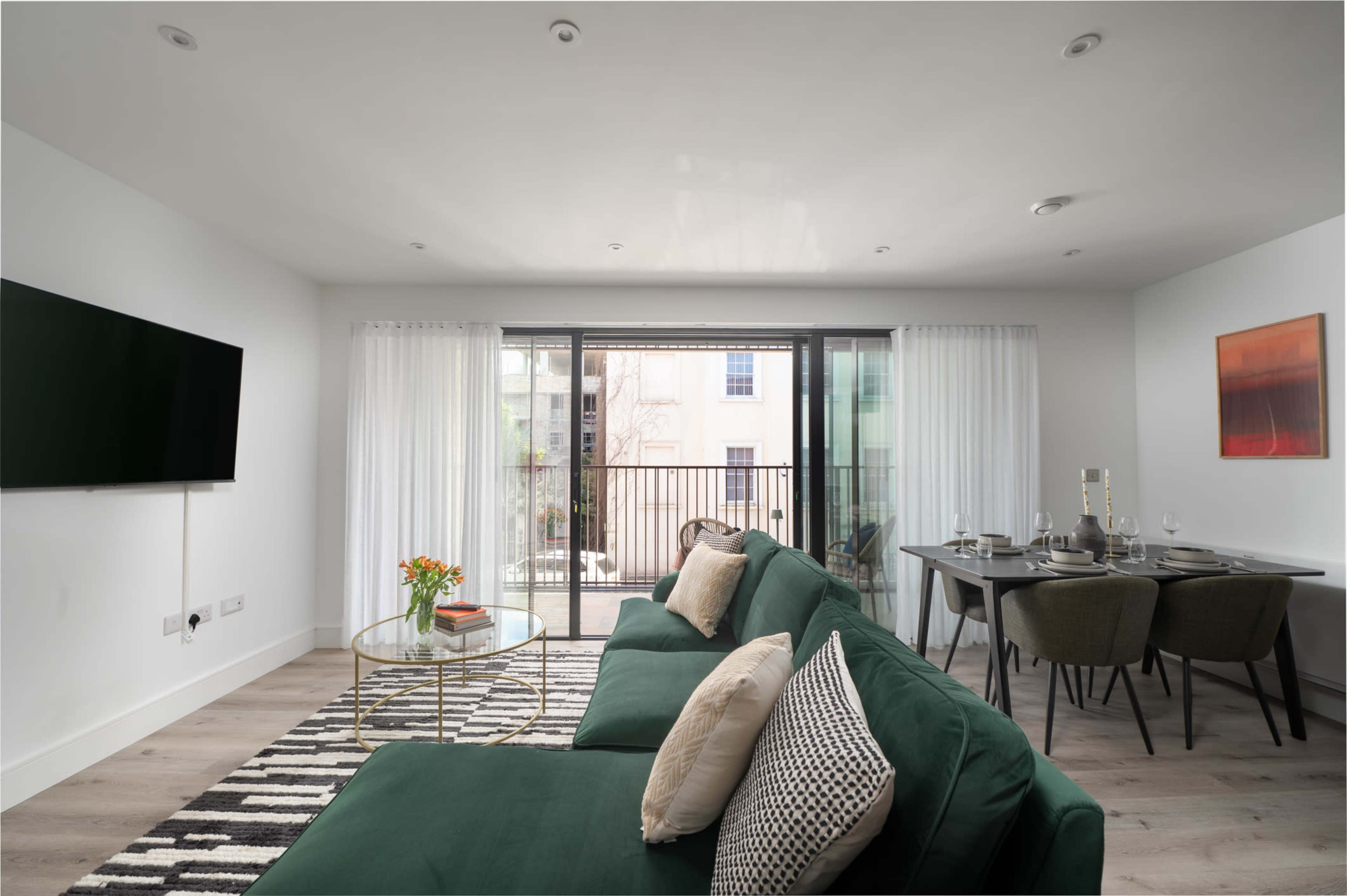 A modern living room features a green sofa, a glass coffee table with a vase of flowers, a dining area with a table set for four, and large sliding glass doors leading to a balcony.