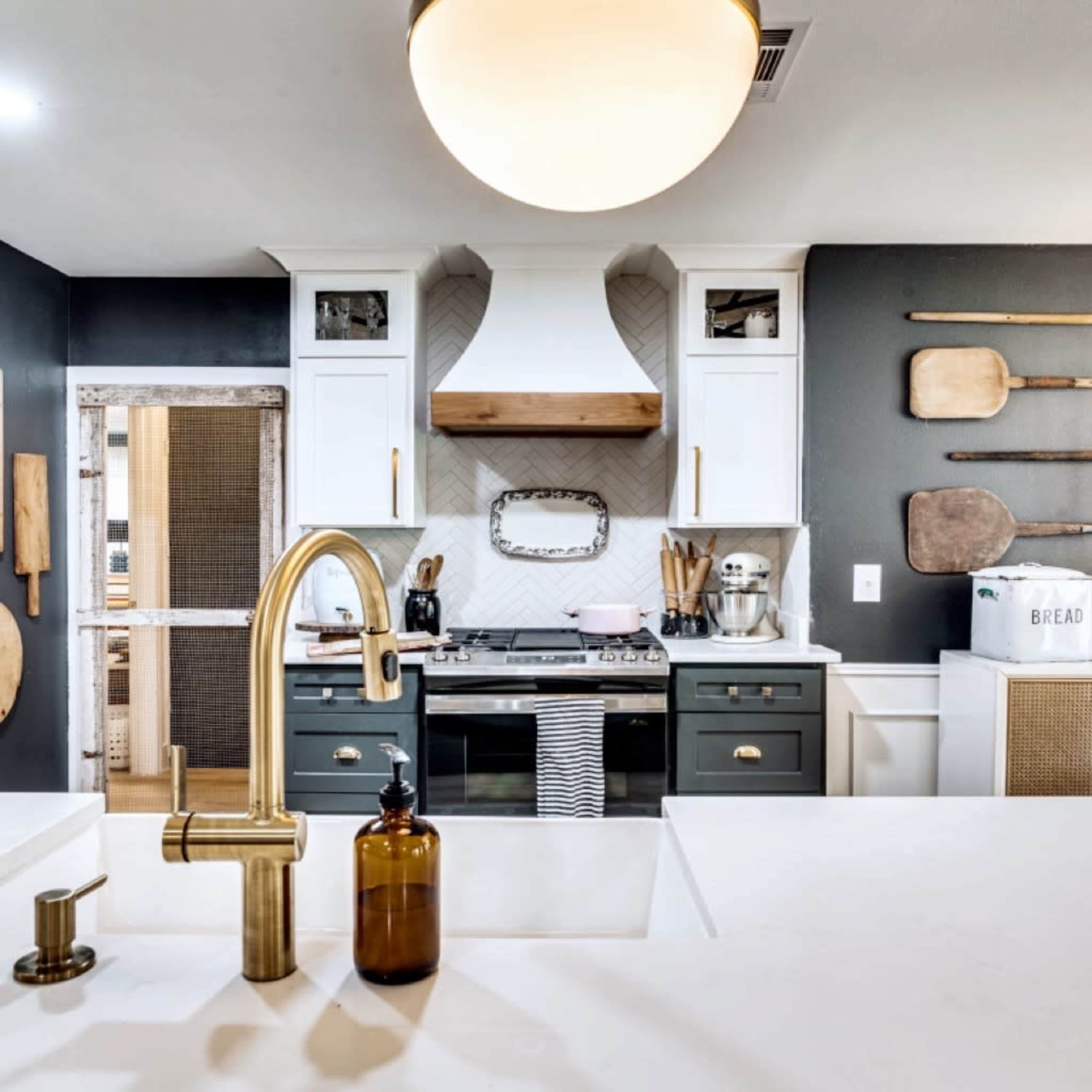 A modern kitchen features a central island with a gold faucet, white cabinetry, a decorative hood above the stove, and wooden utensils displayed on the walls.