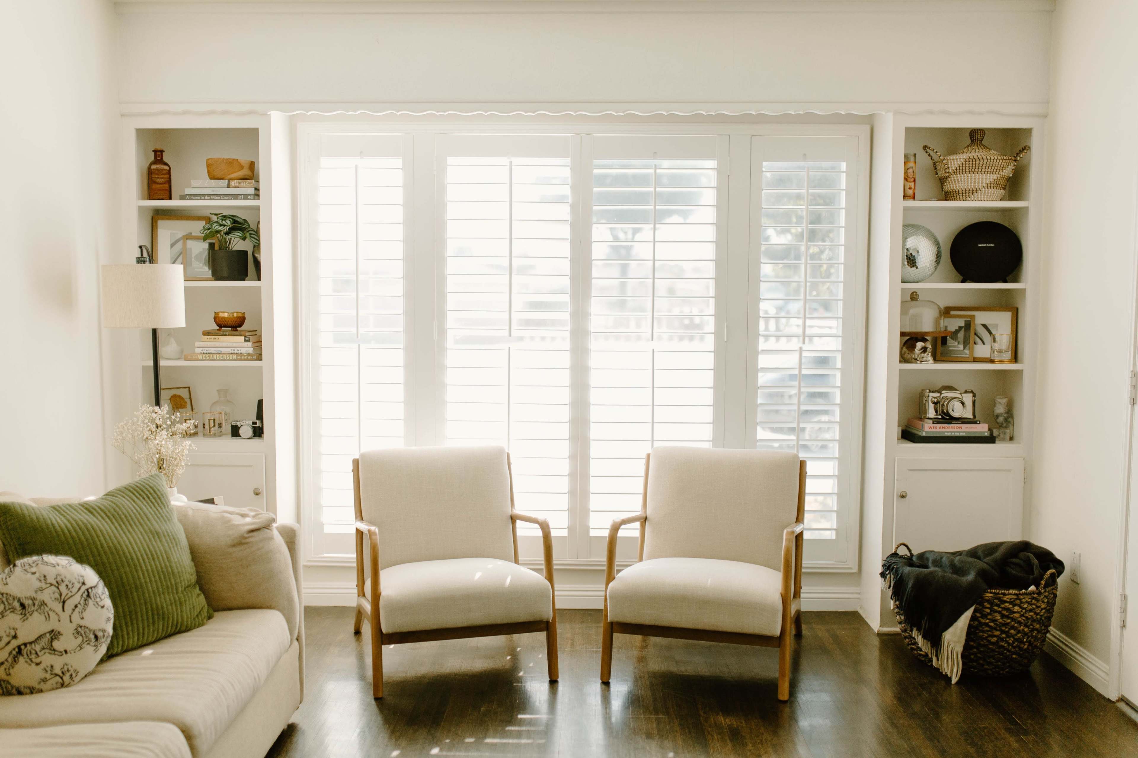 A living room with two cream-colored chairs facing a window covered by white shutters, flanked by built-in shelves displaying various decorative items.
