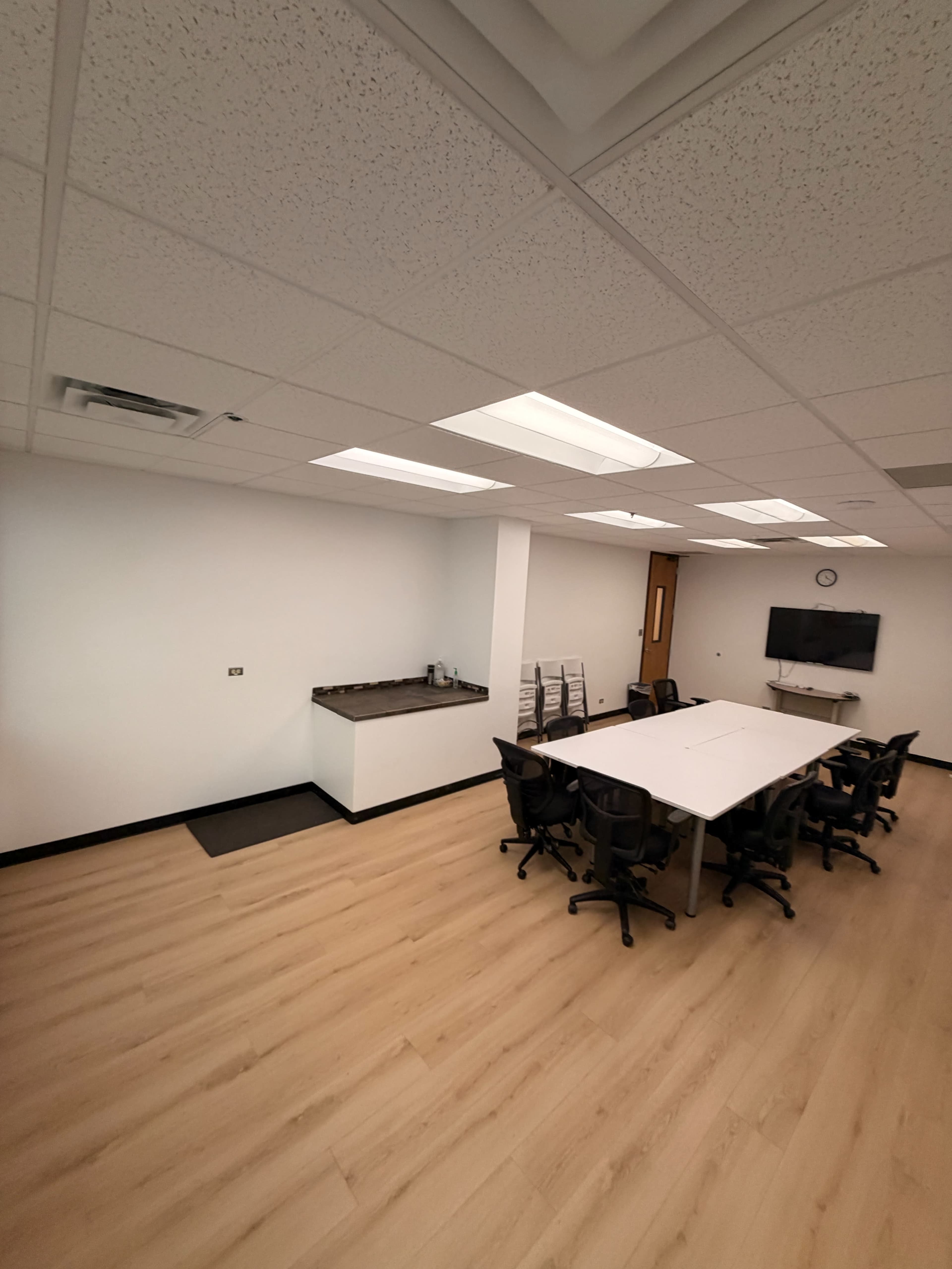 The image shows a modern office space with a large table surrounded by chairs, a kitchenette area on one side, and a wall-mounted television.