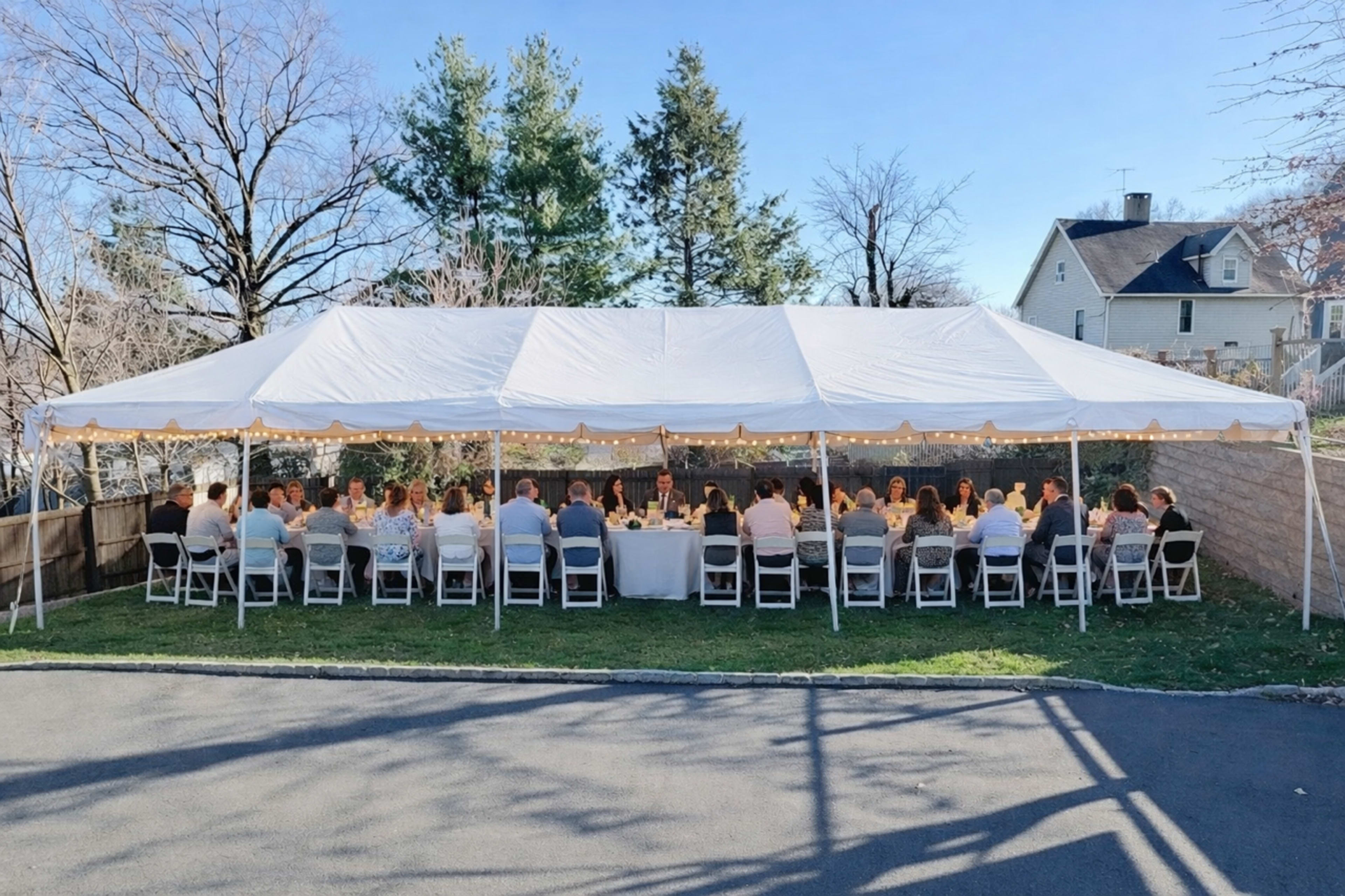 A large group of people is seated at a long table under a white tent in a backyard setting, surrounded by trees and residential houses.