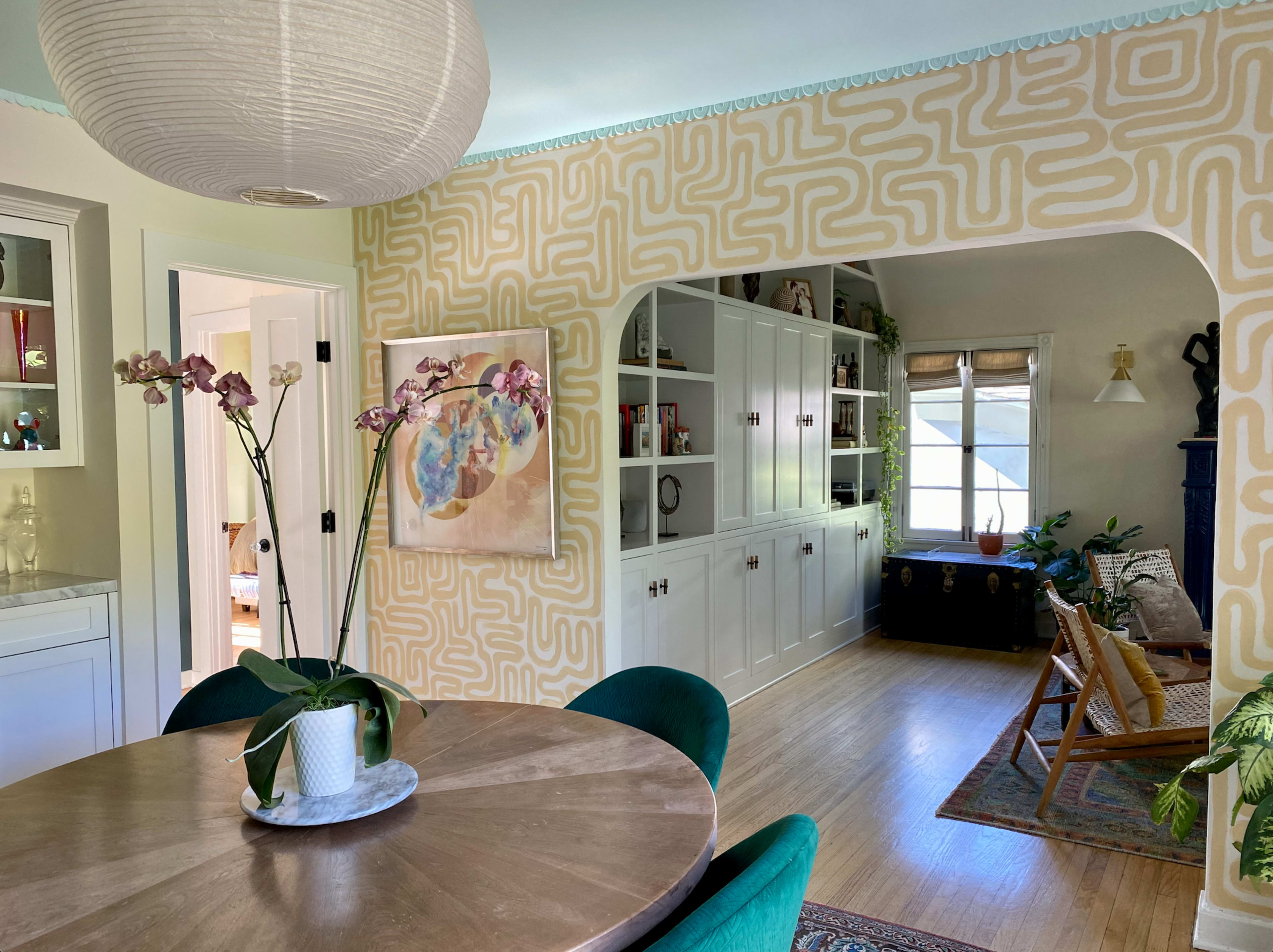 A round table with a plant centerpiece is set in a dining area that opens to a living space featuring bookshelves and large windows.