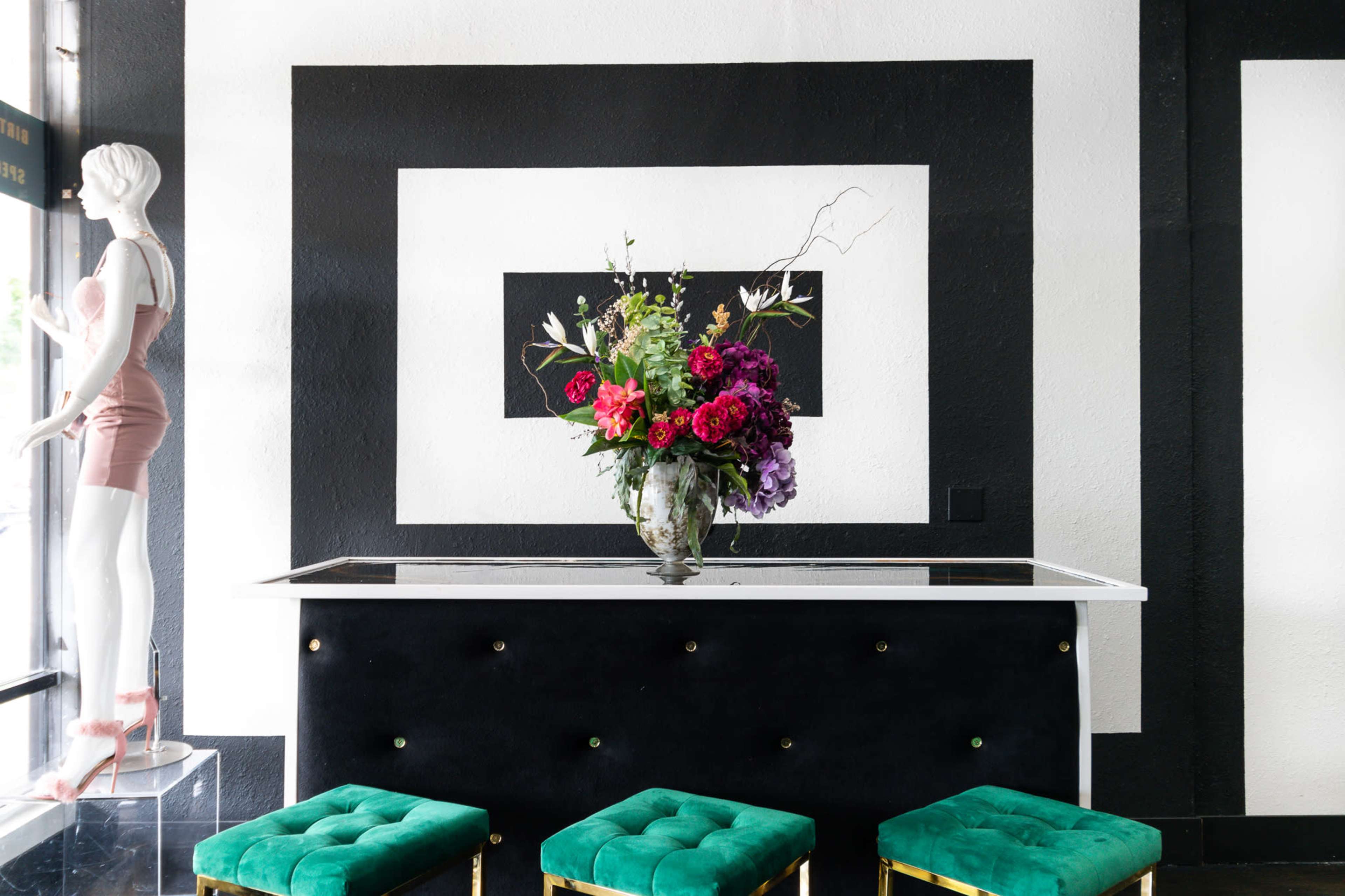A black and white interior features a sleek table adorned with a large floral arrangement, flanked by green stools and a mannequin.