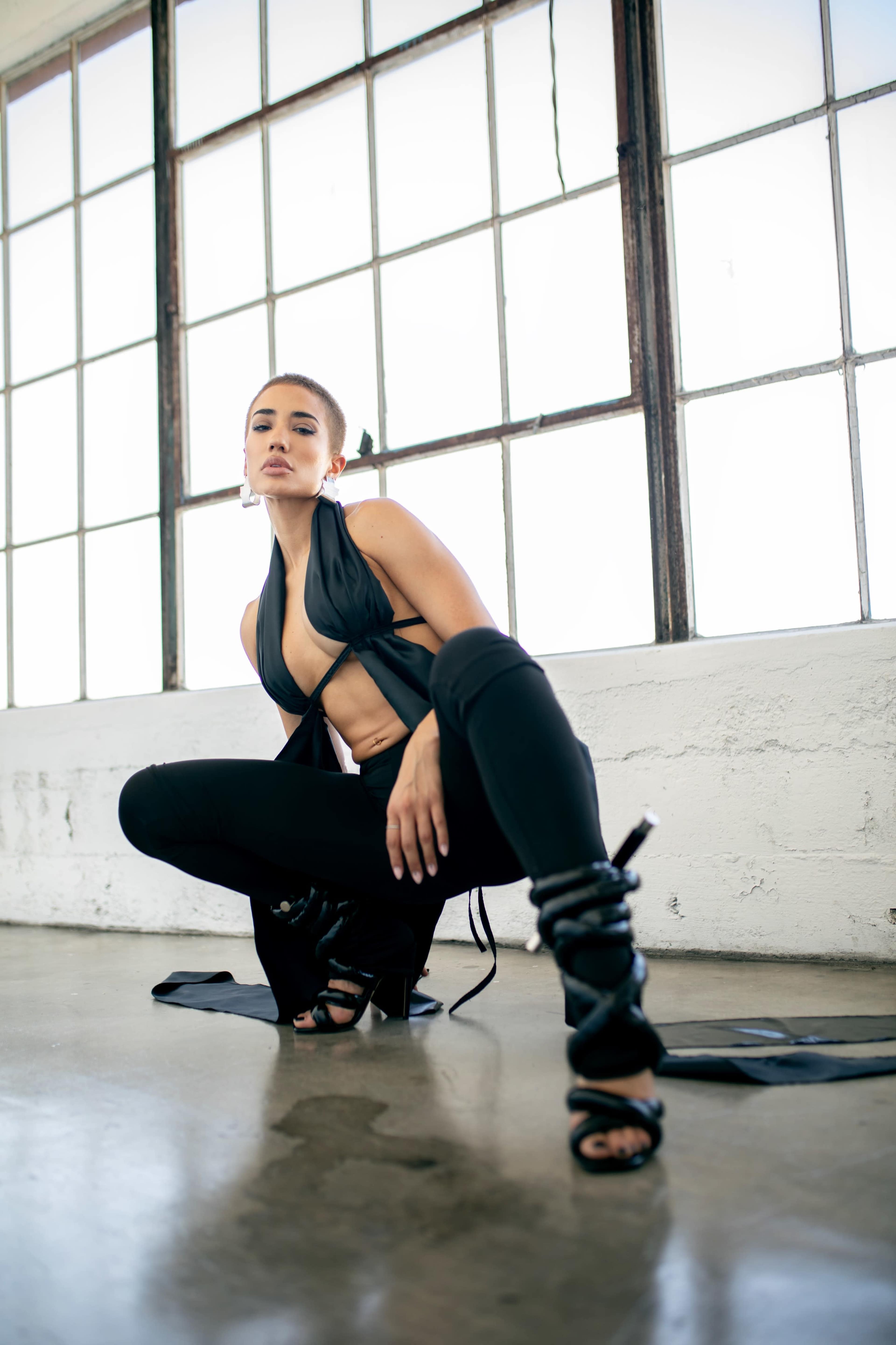 A person poses in a crouched stance wearing a black outfit and strappy heels, against a backdrop of large windows.