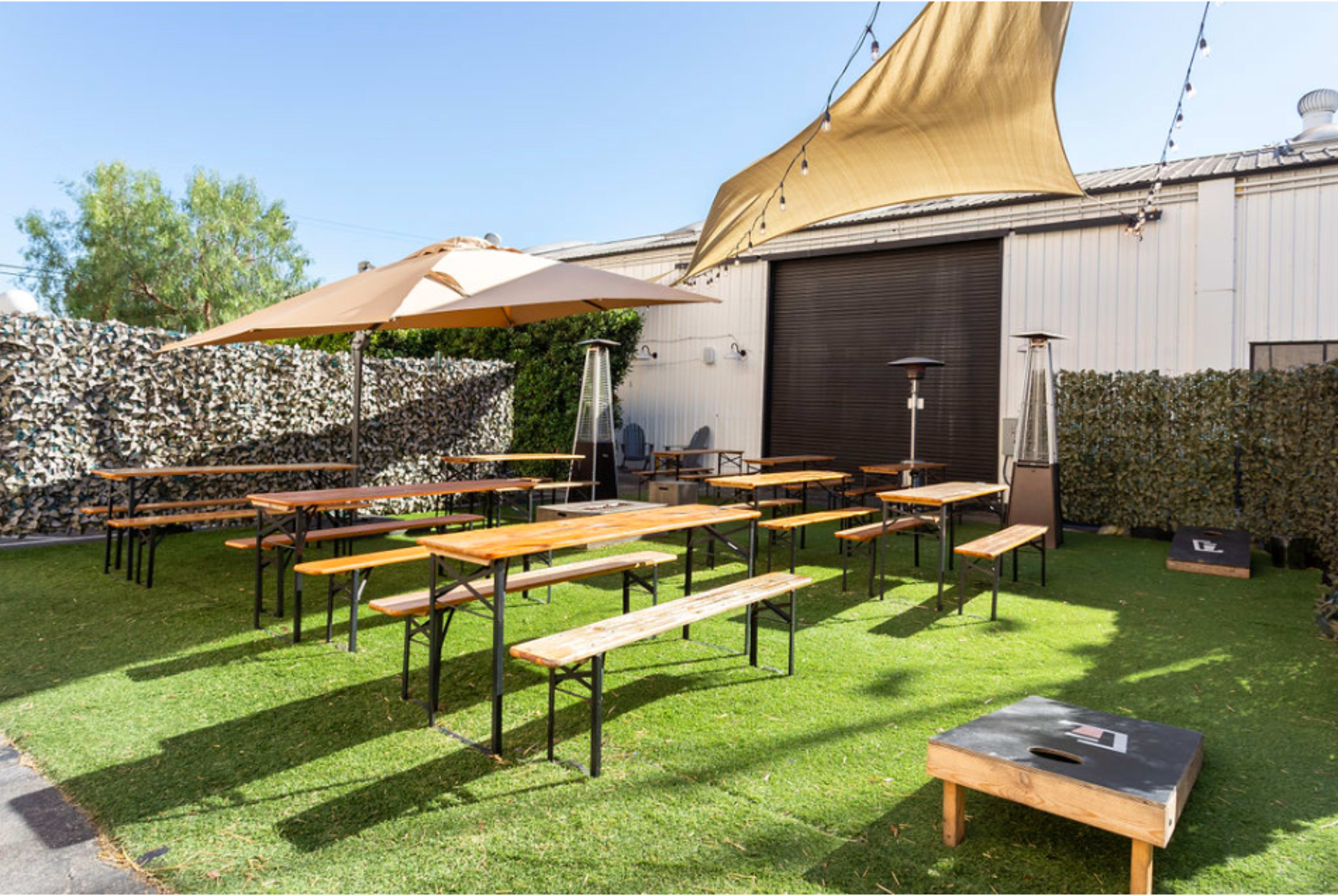 The image shows an outdoor seating area with several wooden picnic tables and benches, shaded by large umbrellas and surrounded by decorative foliage.