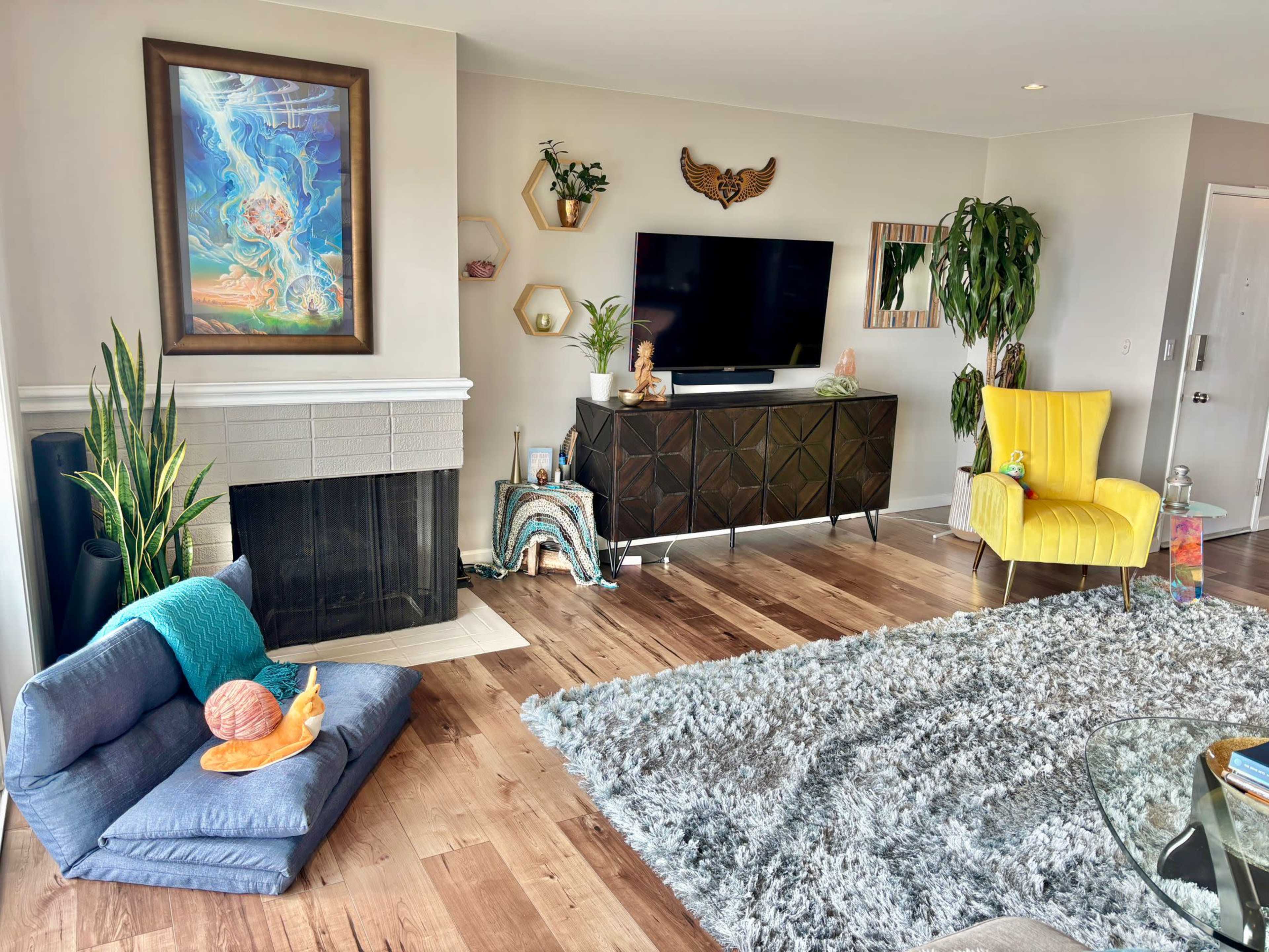 The room features a modern living space with a gray couch, a yellow chair, a television mounted on the wall, and decorative plants.