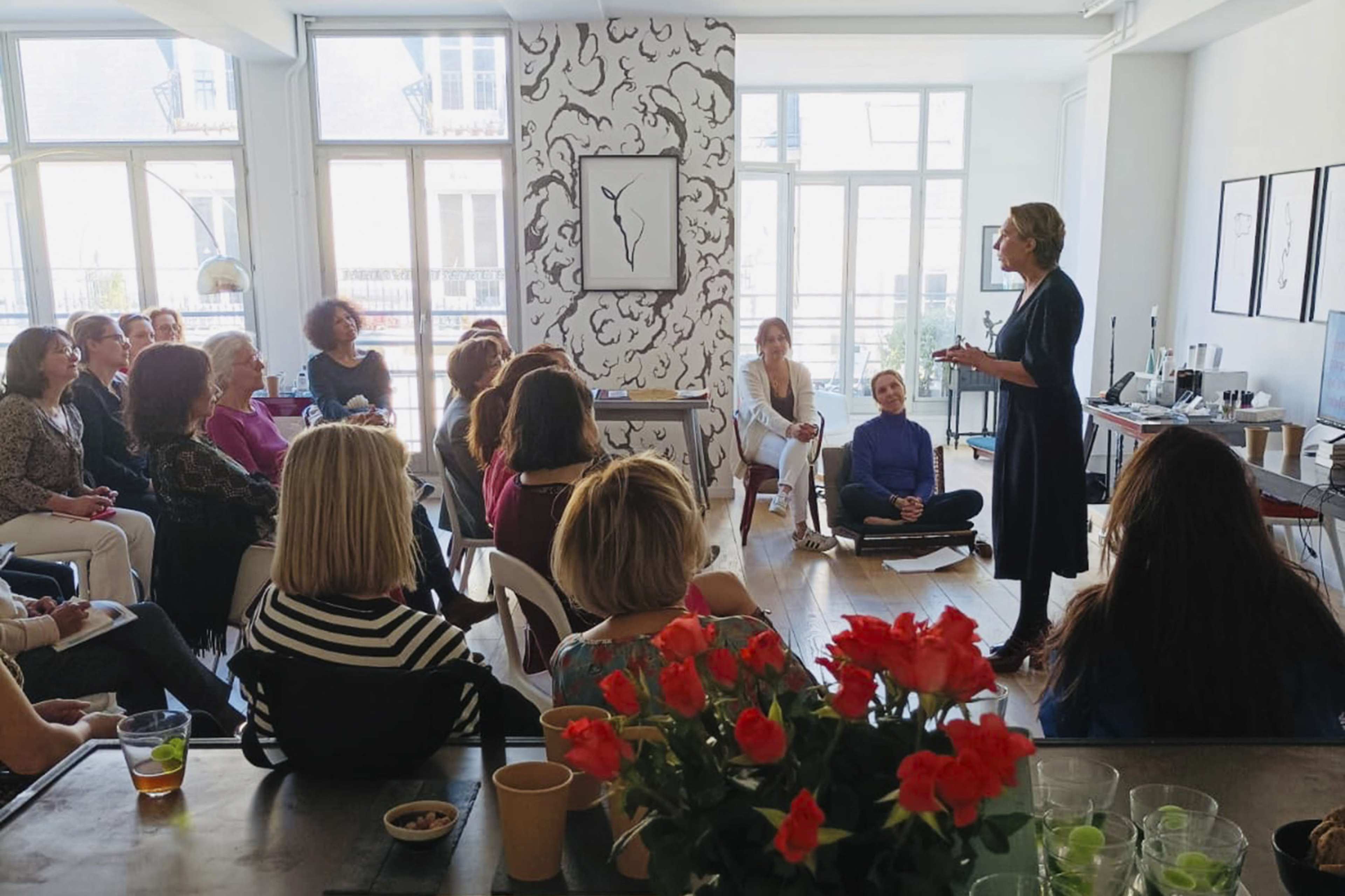 A speaker addresses an audience of seated women in a bright, spacious room adorned with artwork and plants.