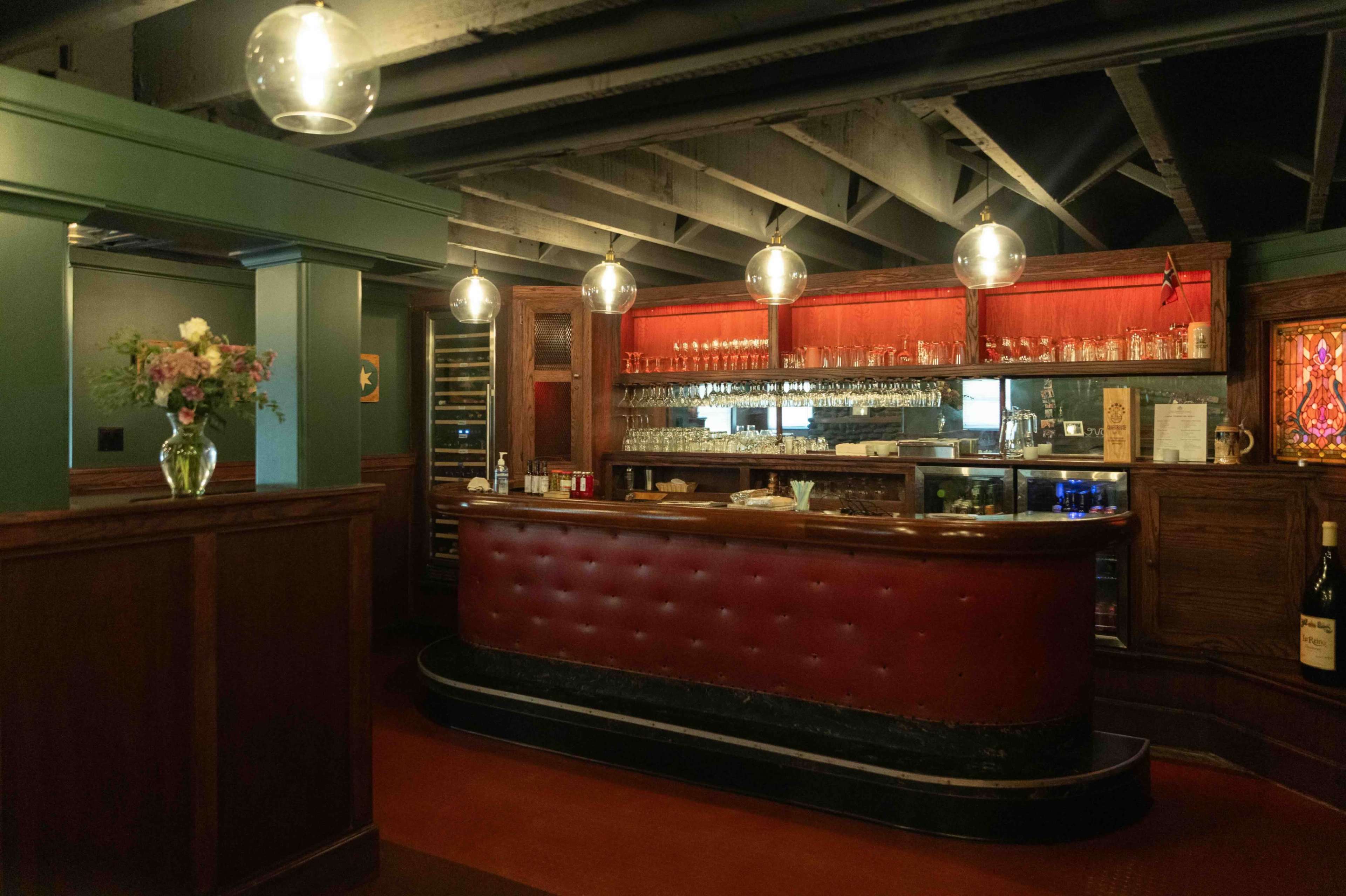 The image shows a vintage-style bar with a red leather-faced counter, glassware displayed on shelves behind it, and warm pendant lights illuminating the space.