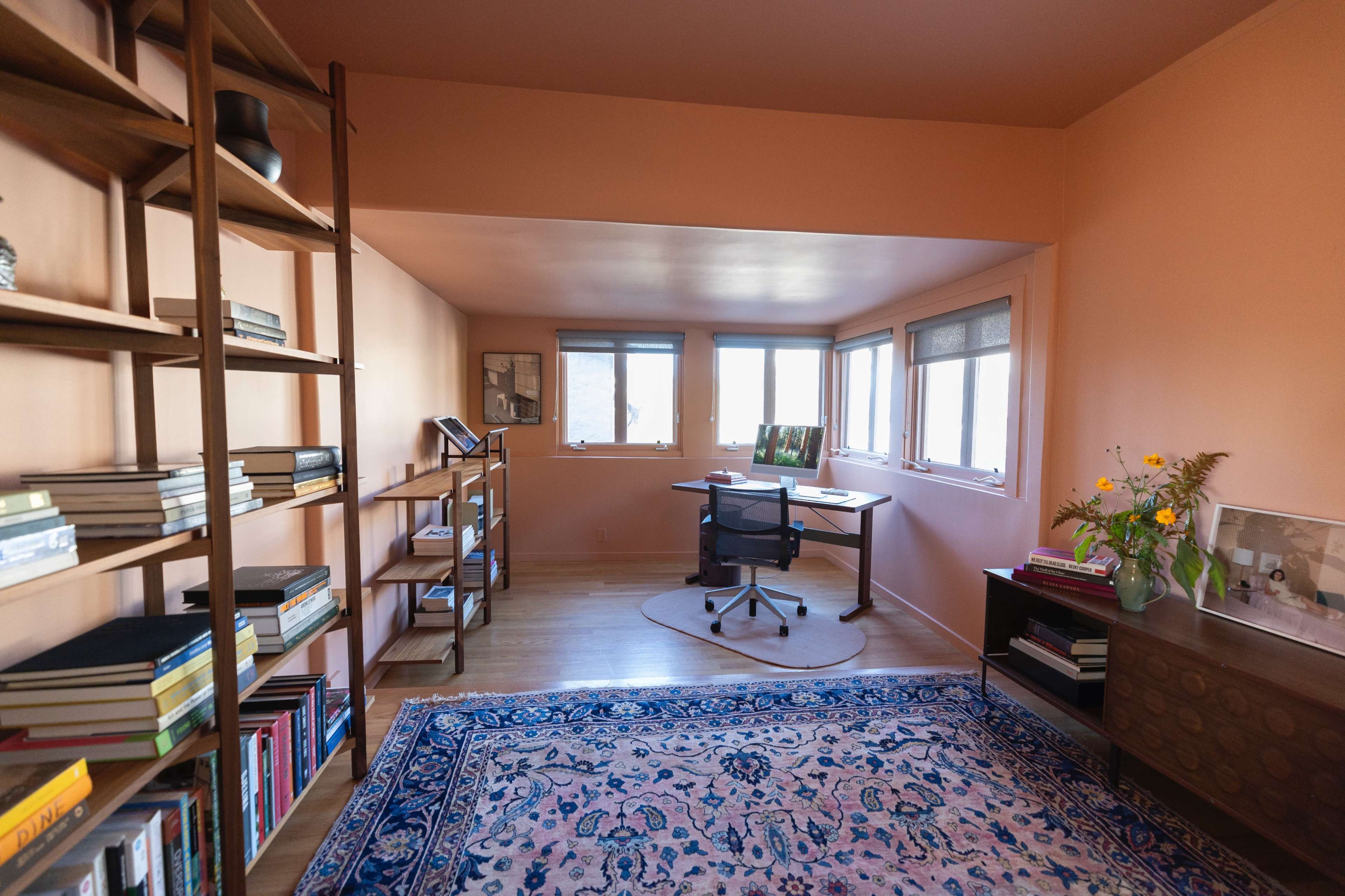 The image shows a brightly colored, minimalist room with a wooden desk, a rolling chair, large windows, bookshelves filled with books, and a Persian-style rug.