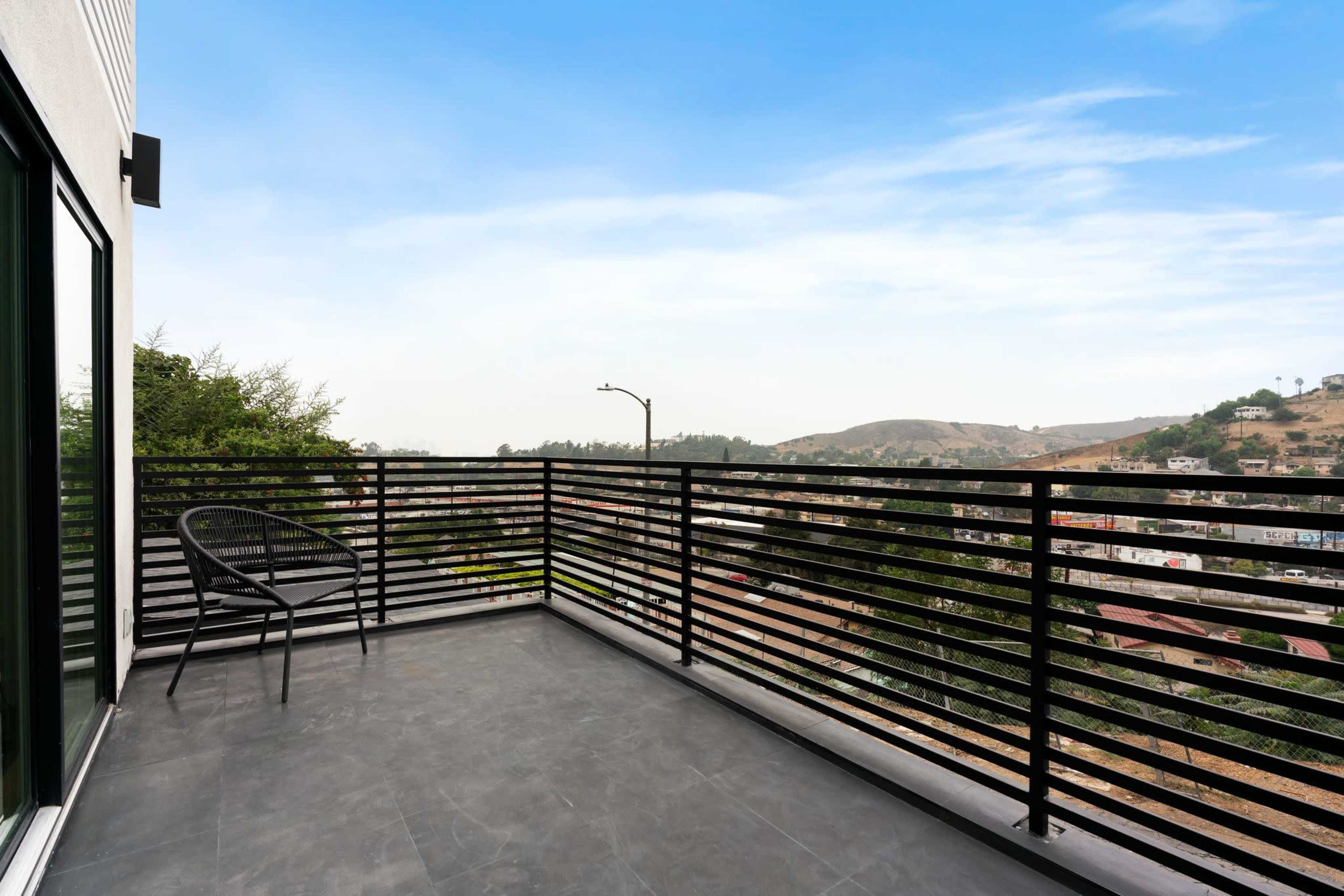 A balcony with black metal railing overlooks a hilly landscape and residential area under a clear sky.
