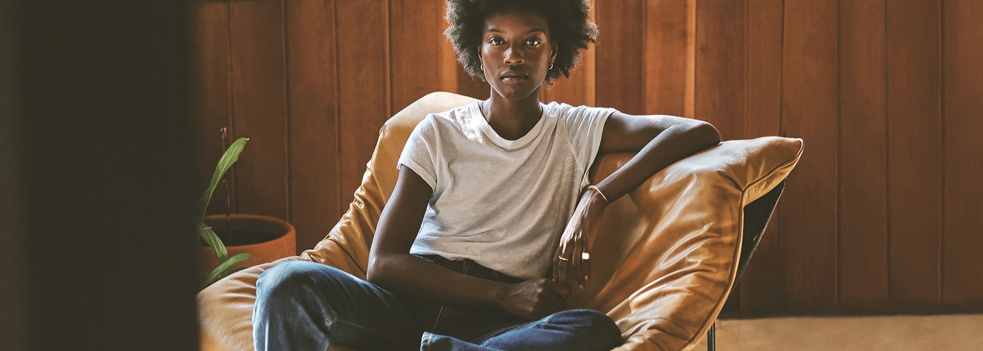 A person sits on a large, comfortable chair in a room with wooden walls, wearing a plain white t-shirt and blue jeans.