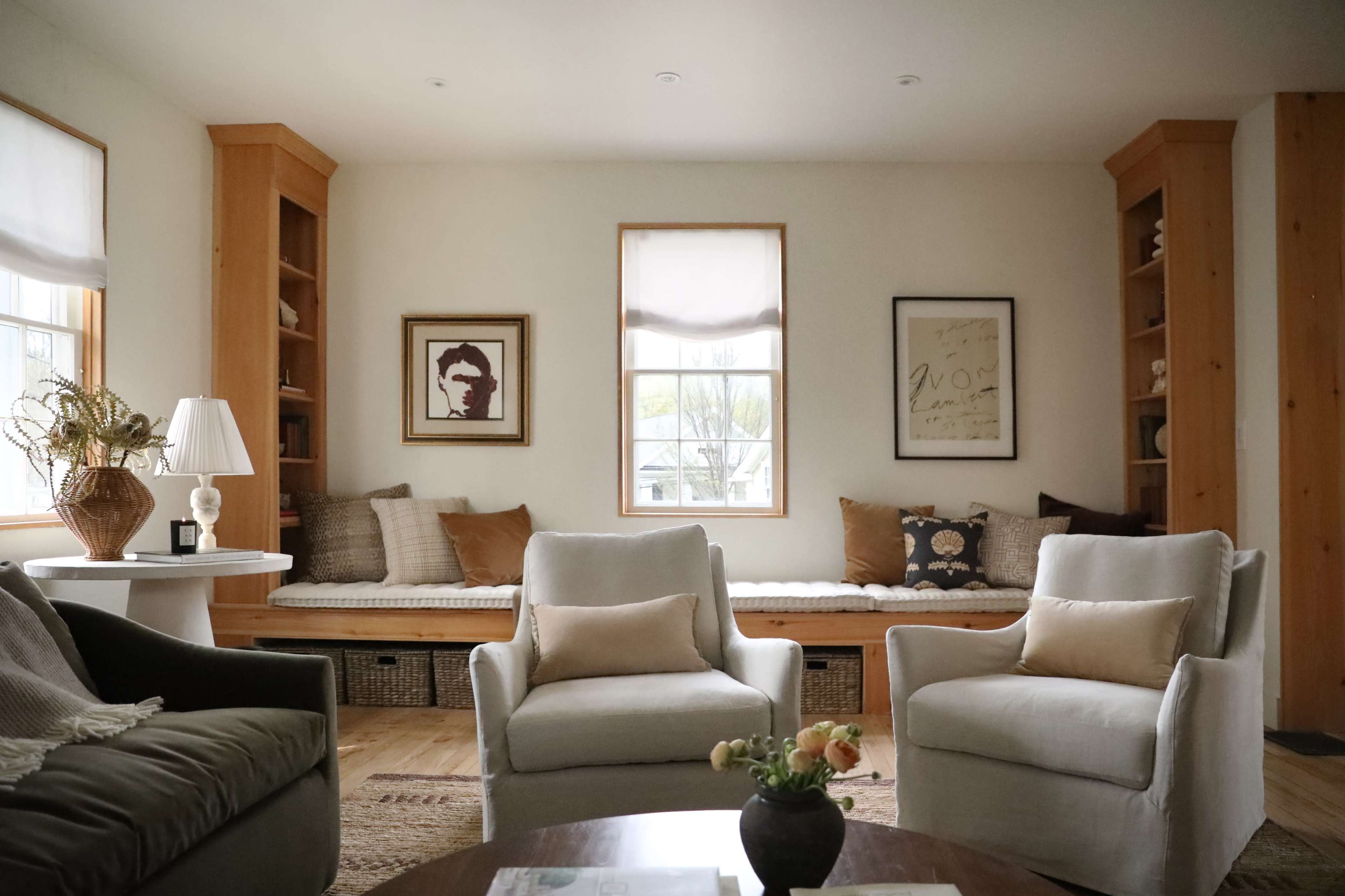 A cozy living room features two upholstered armchairs facing a round coffee table, with large windows and a built-in bench adorned with cushions against the wall.