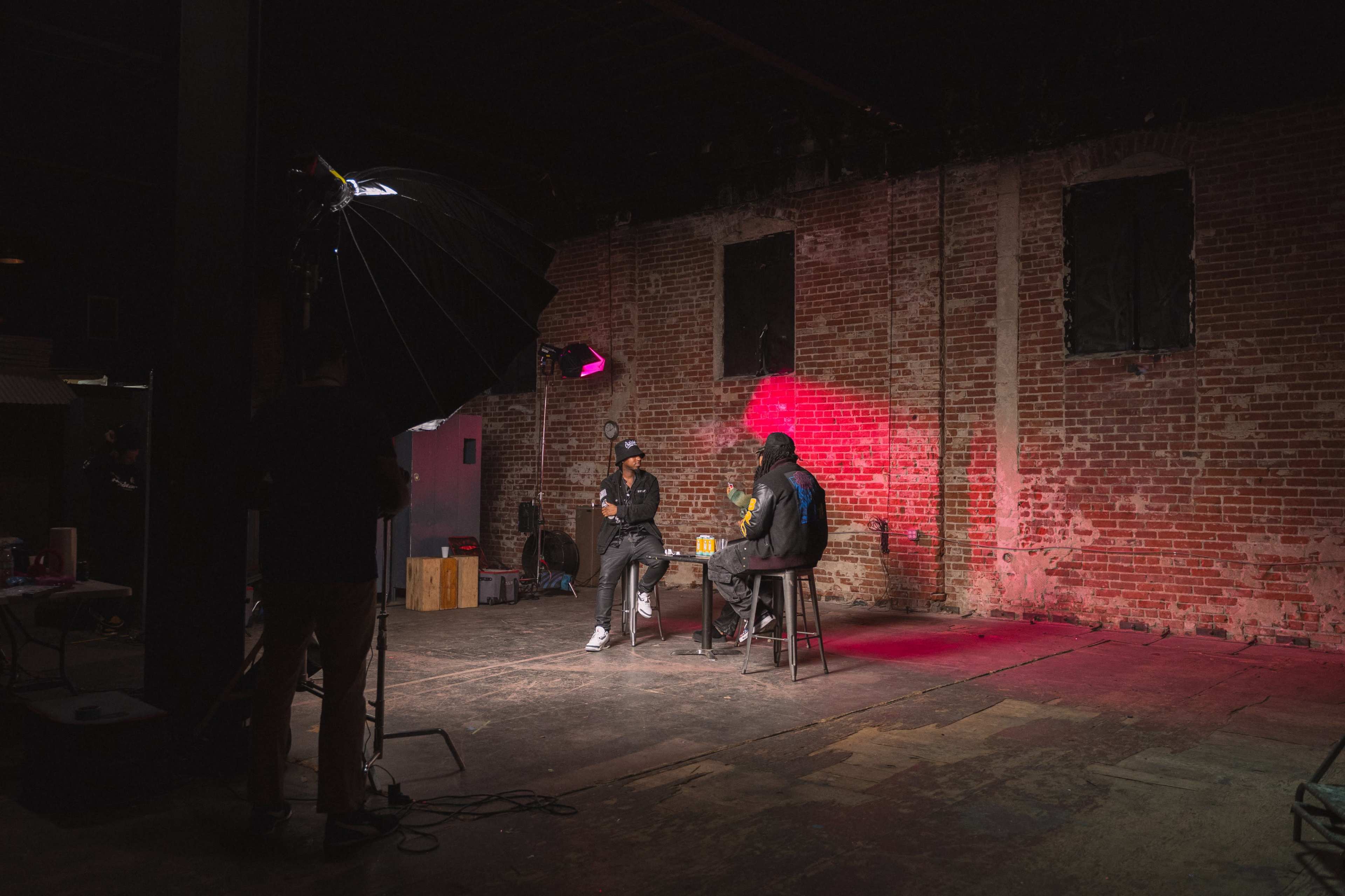 Two individuals sit on stools in a poorly lit room with exposed brick walls, while a large softbox light illuminates the scene.