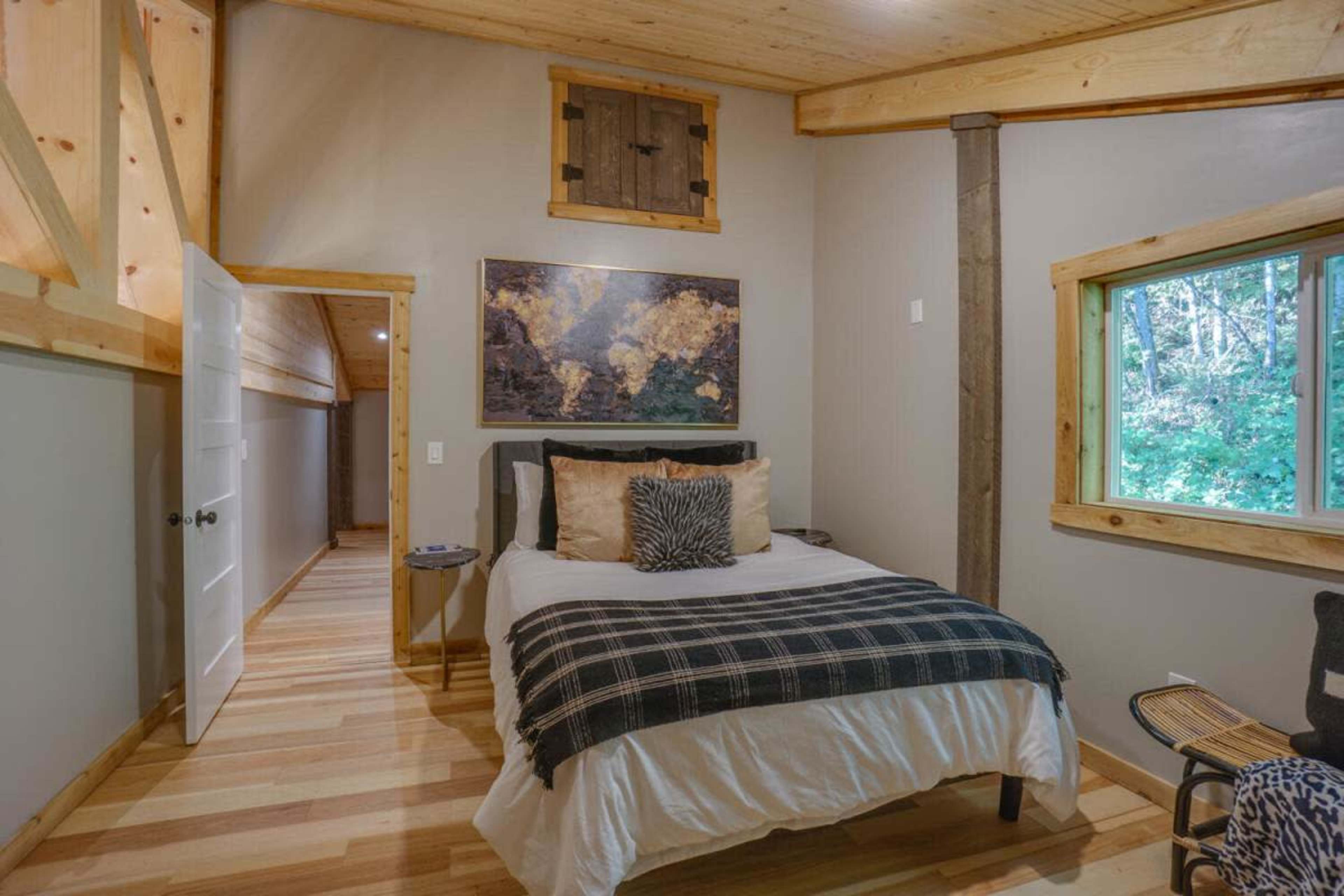 A small bedroom features a bed with a plaid throw, a world map on the wall, and a window overlooking a wooded area.
