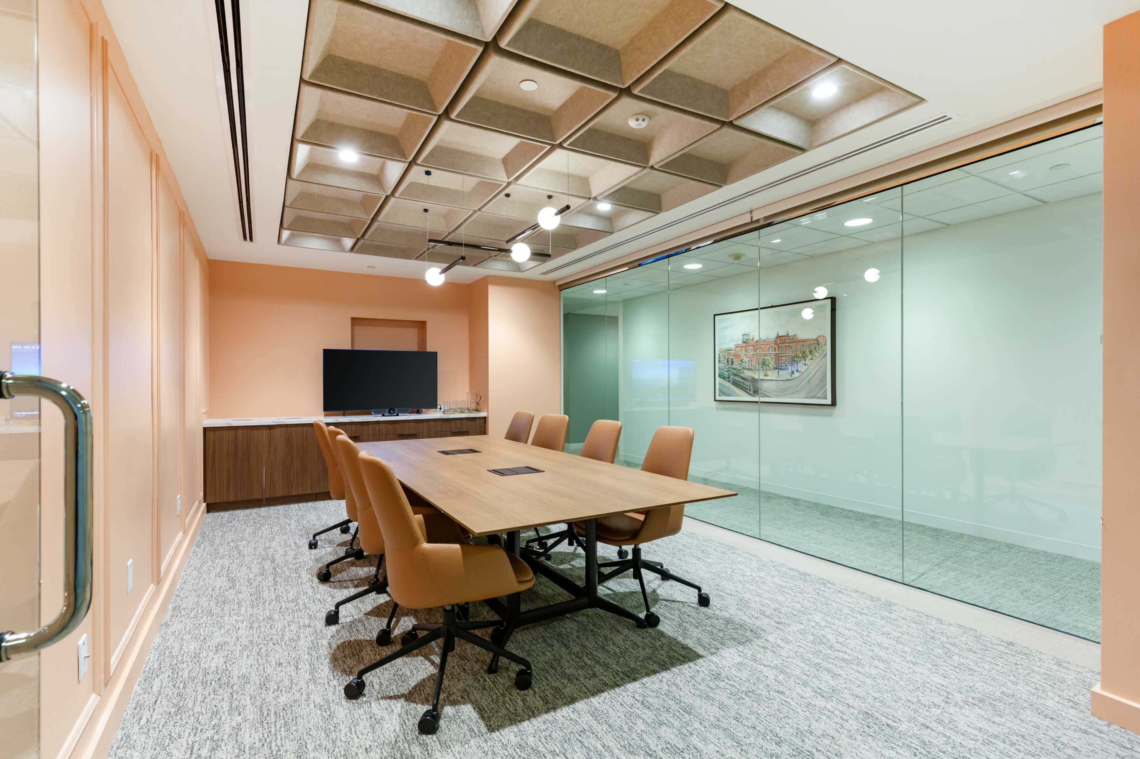A modern conference room features a large wooden table surrounded by ergonomic chairs, with a wall-mounted screen and a glass wall allowing visibility into the next space.