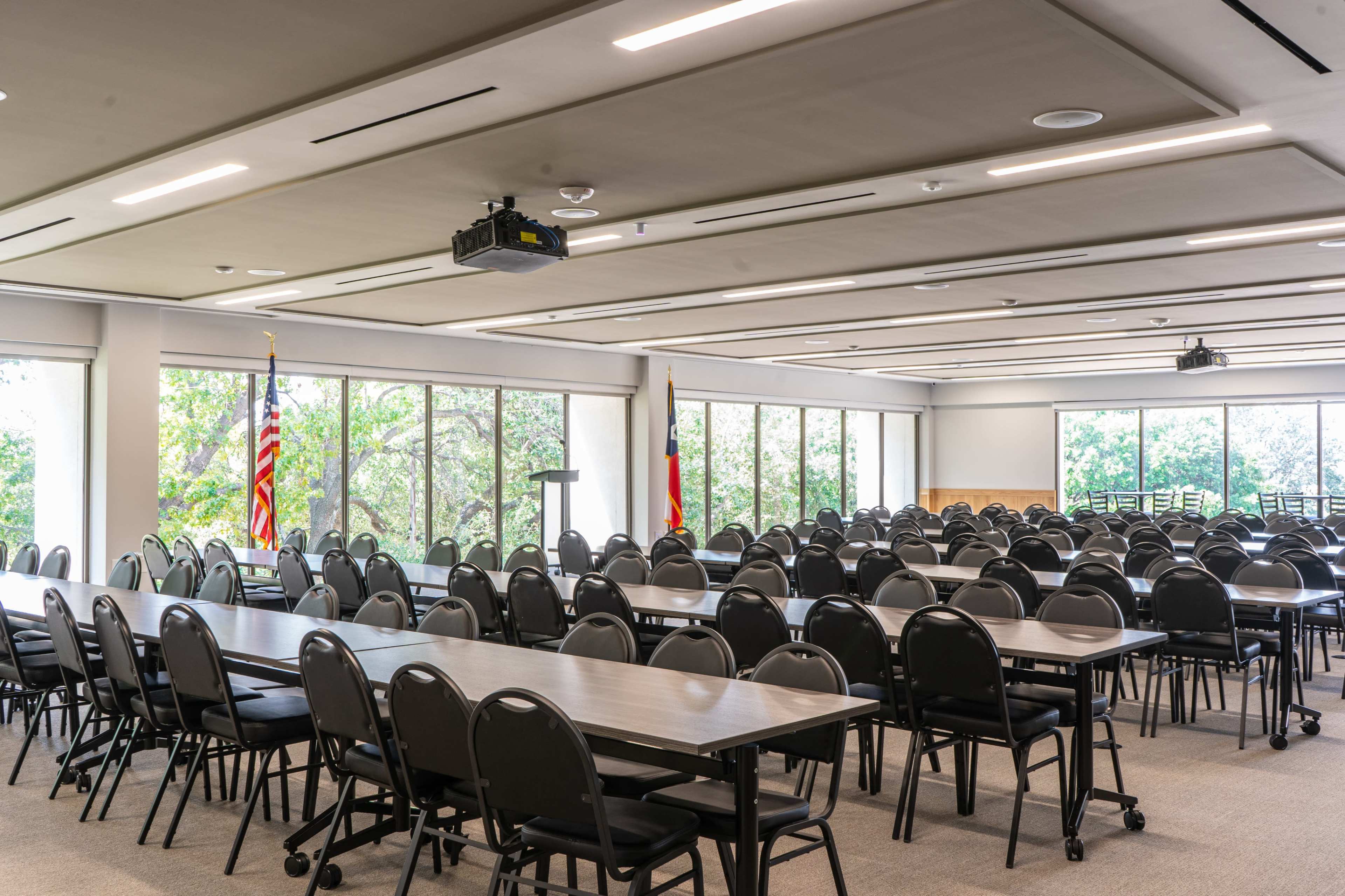 A large, empty conference room features multiple rows of tables and chairs, with American and state flags displayed near the windows.