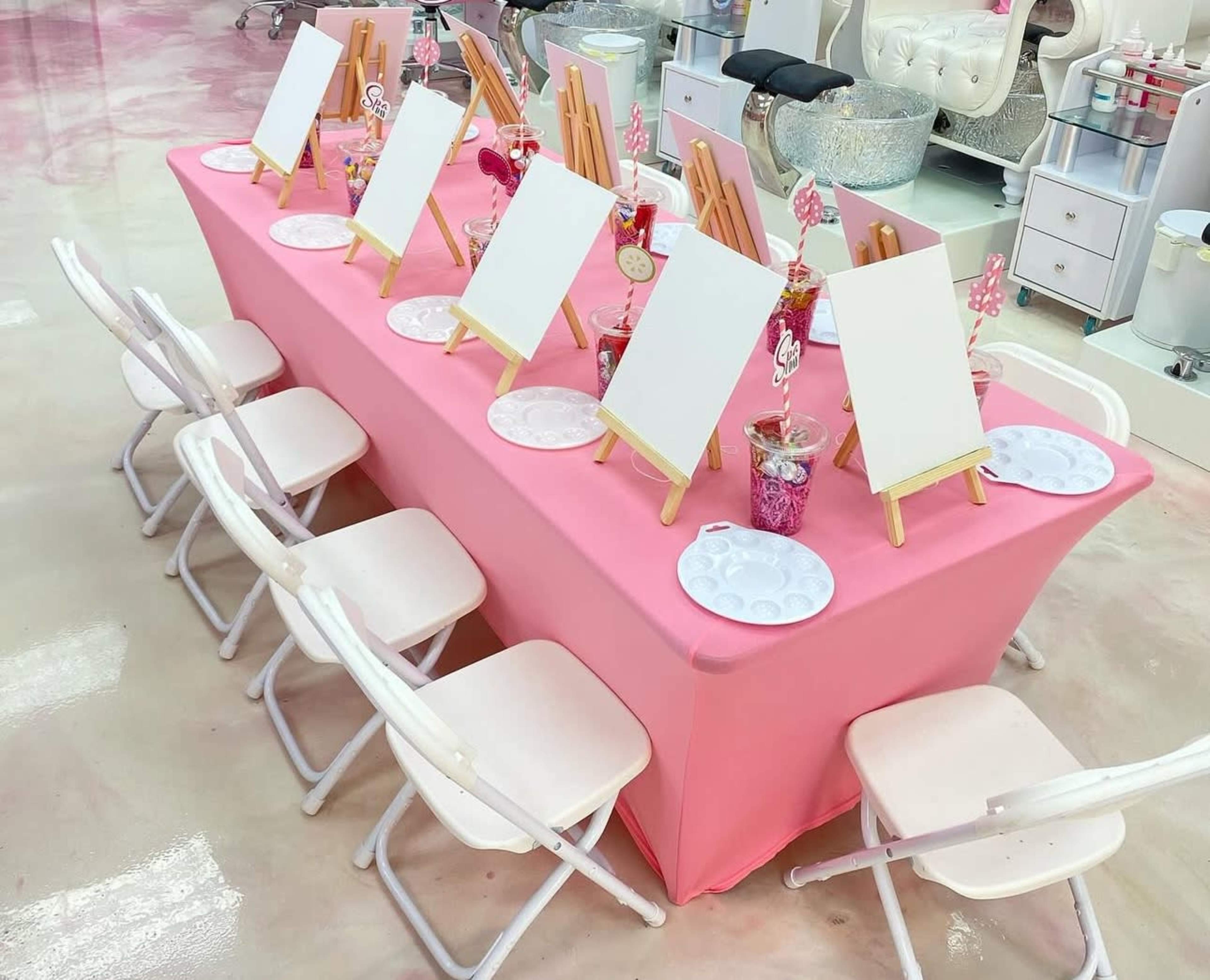 A long pink table is set with blank canvases on easels, surrounded by white folding chairs, in a brightly lit room.
