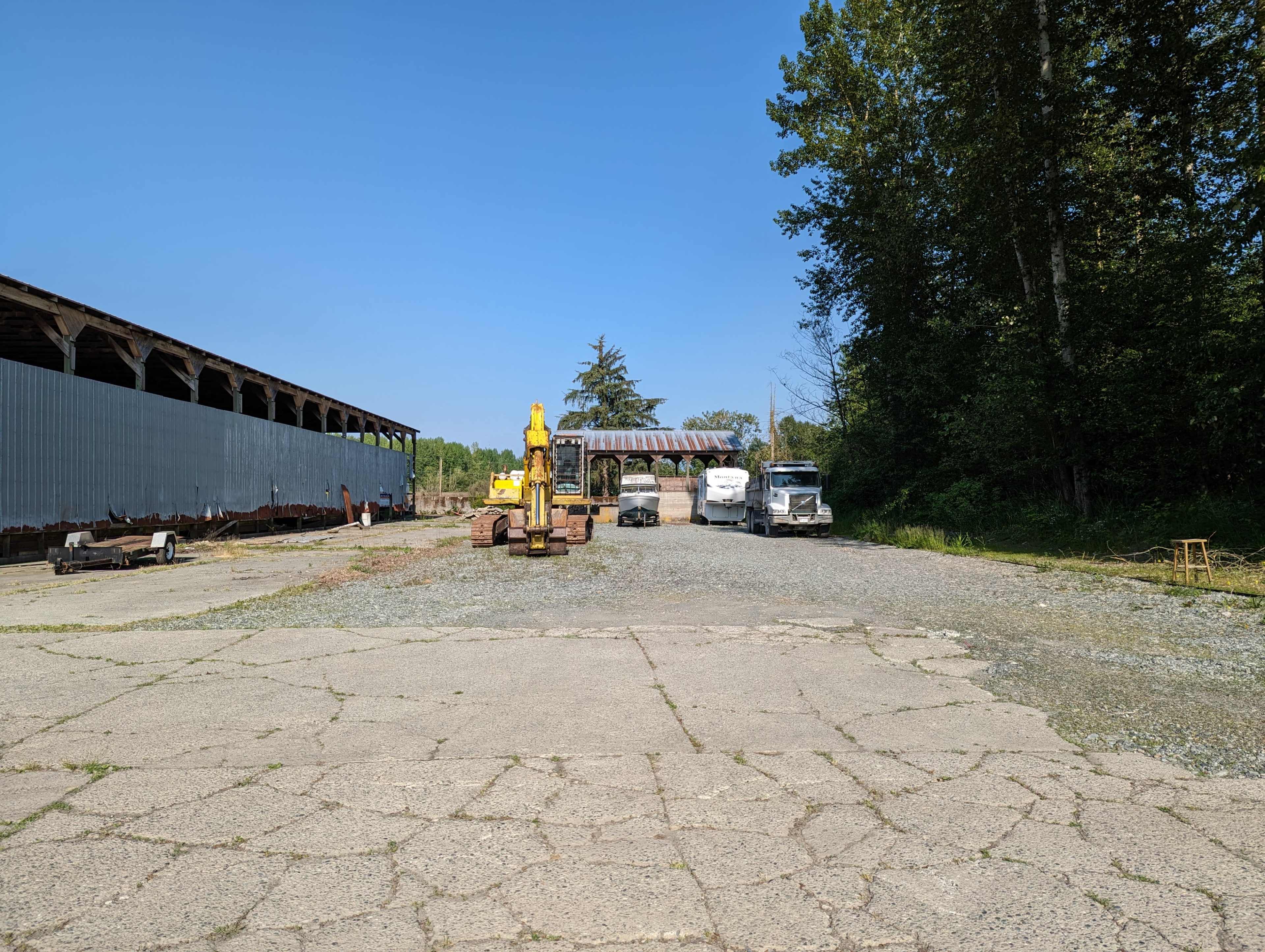 A gravel lot features heavy machinery and parked trucks, bordered by a metal building and trees under a clear blue sky.