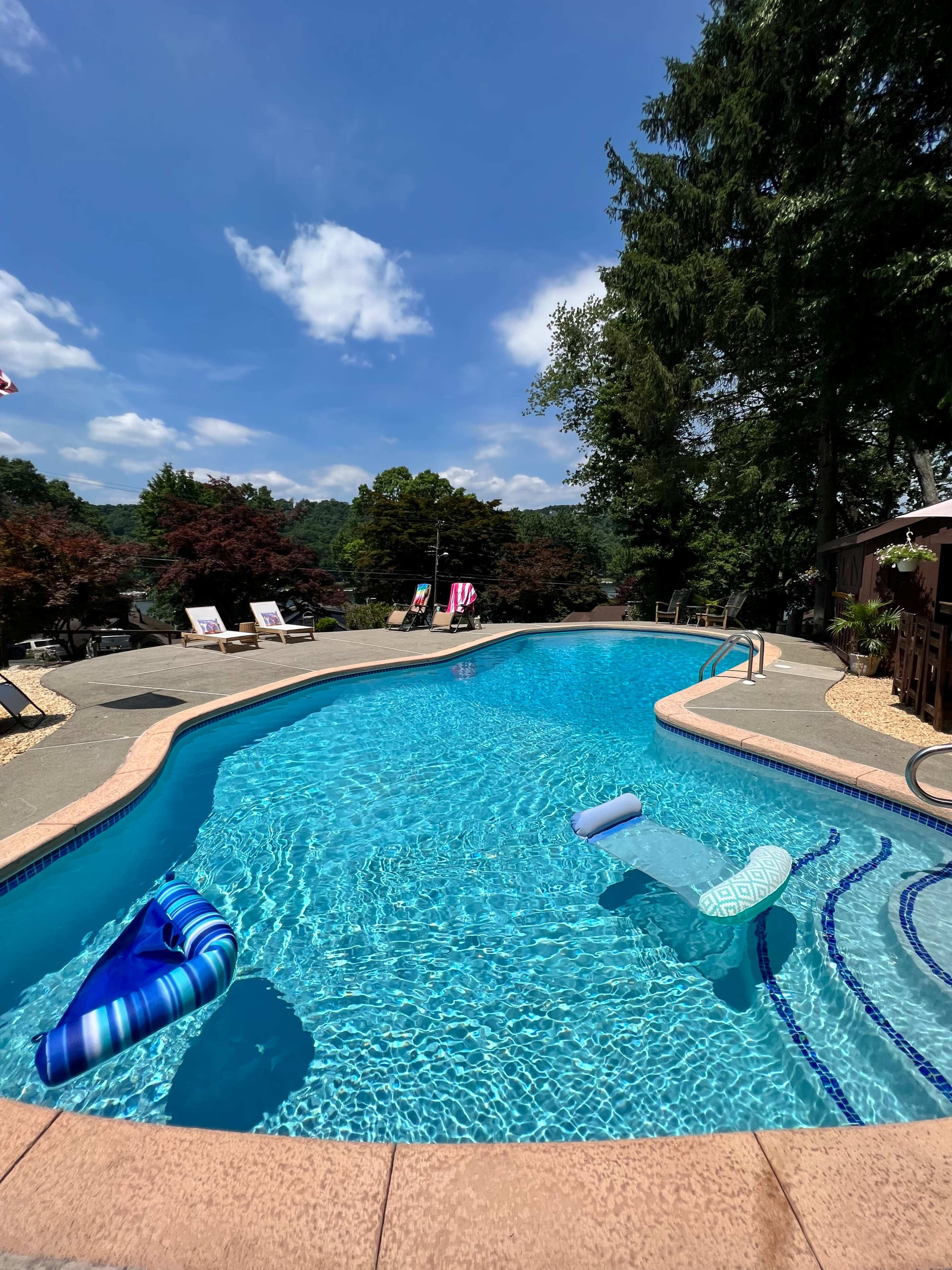 A clear swimming pool surrounded by trees and lounge chairs on a sunny day.