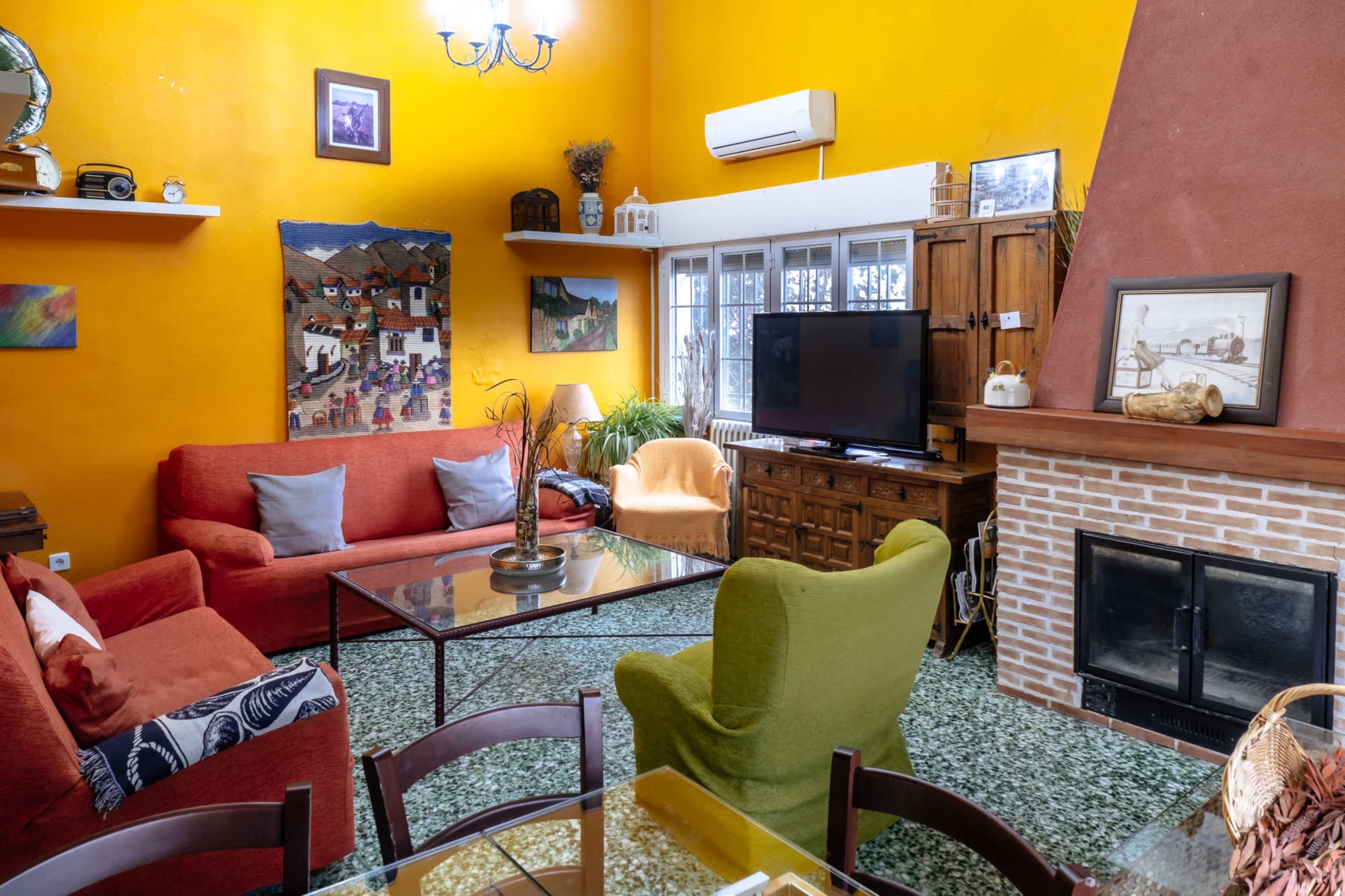 The scene is a vibrant living room featuring orange walls, various seating options, a television, a fireplace, and a colorful painting.