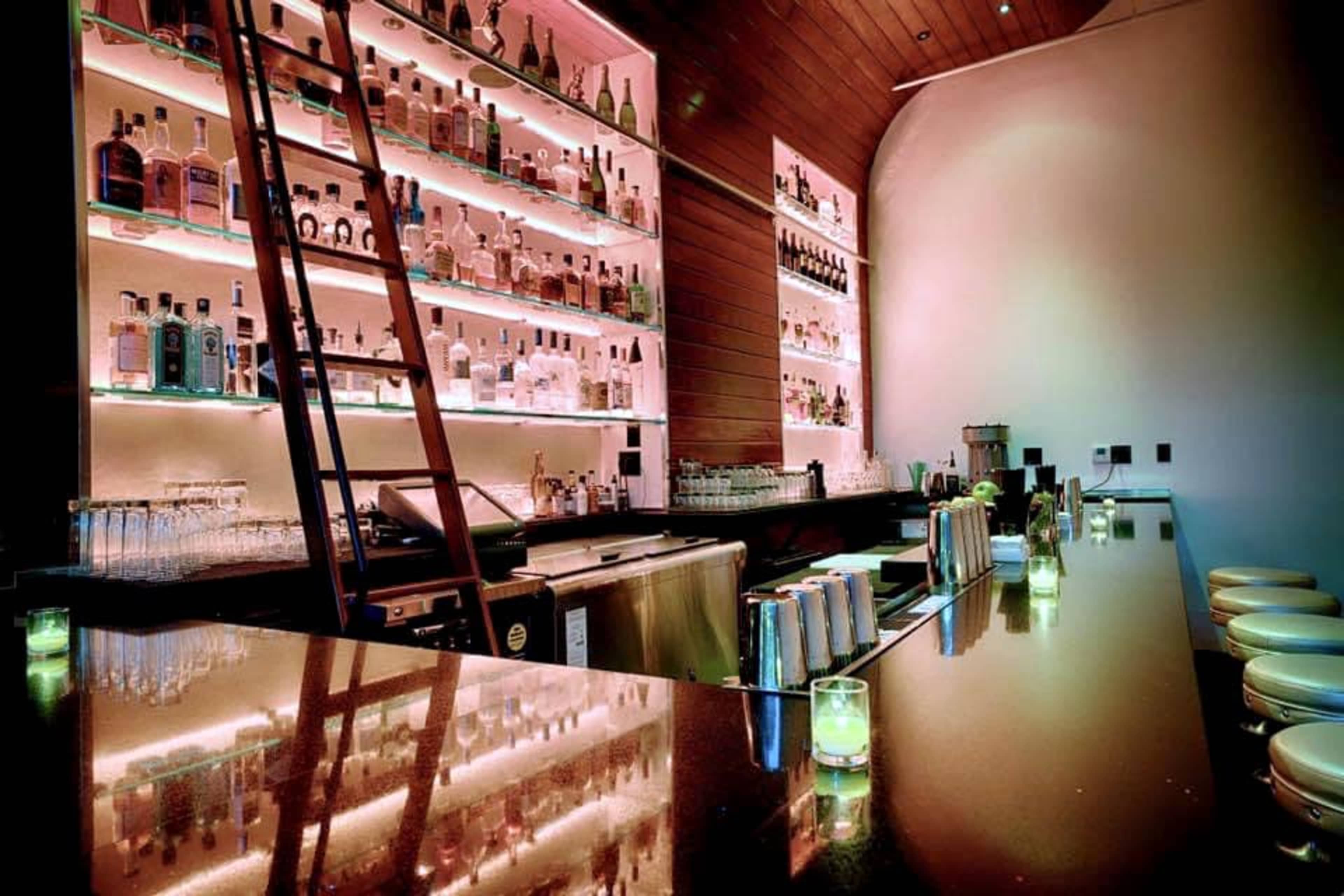 A modern bar features illuminated shelves stocked with various bottles, a polished countertop, and bar stools.