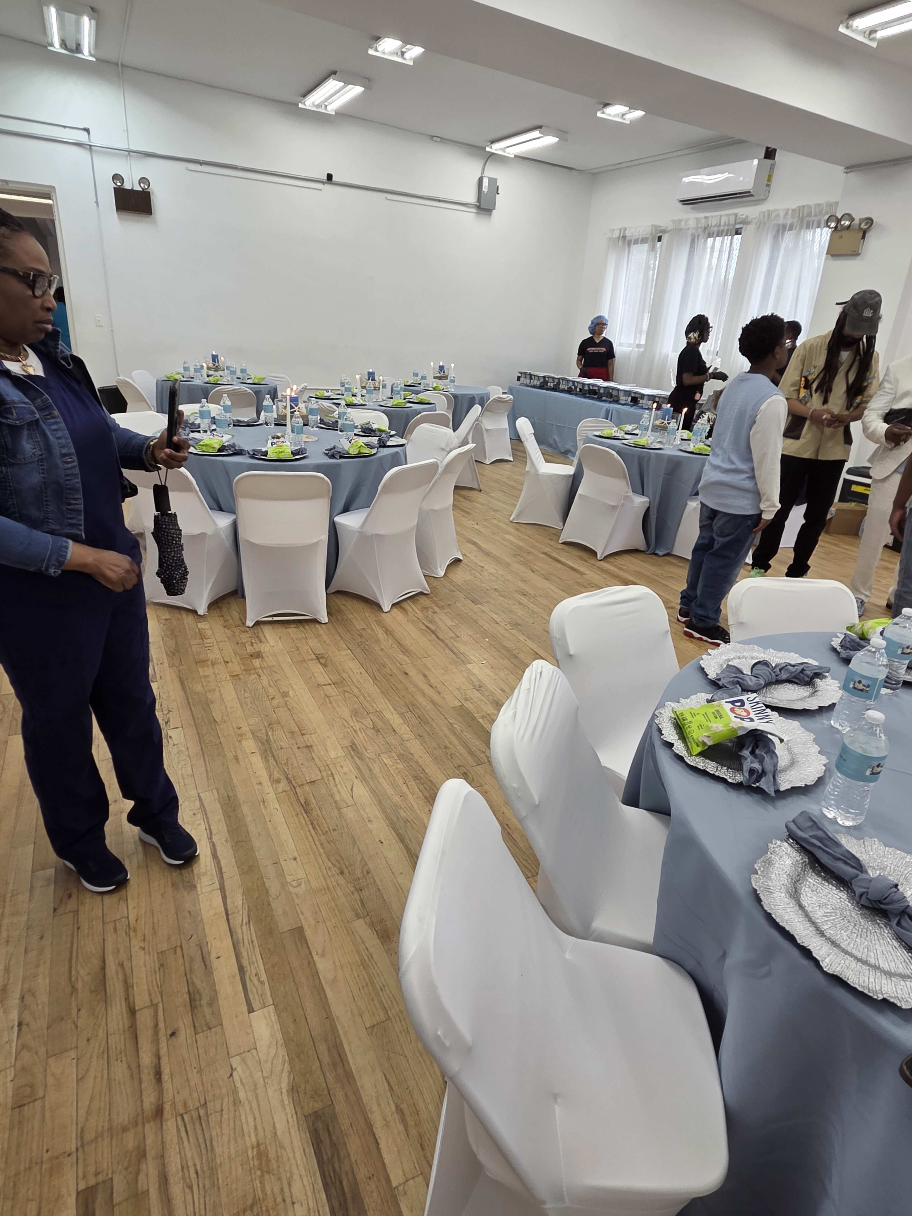 The image shows a banquet-style room arranged with tables set for an event, featuring gray table covers, chairs, and various decorations, while individuals stand around the space.