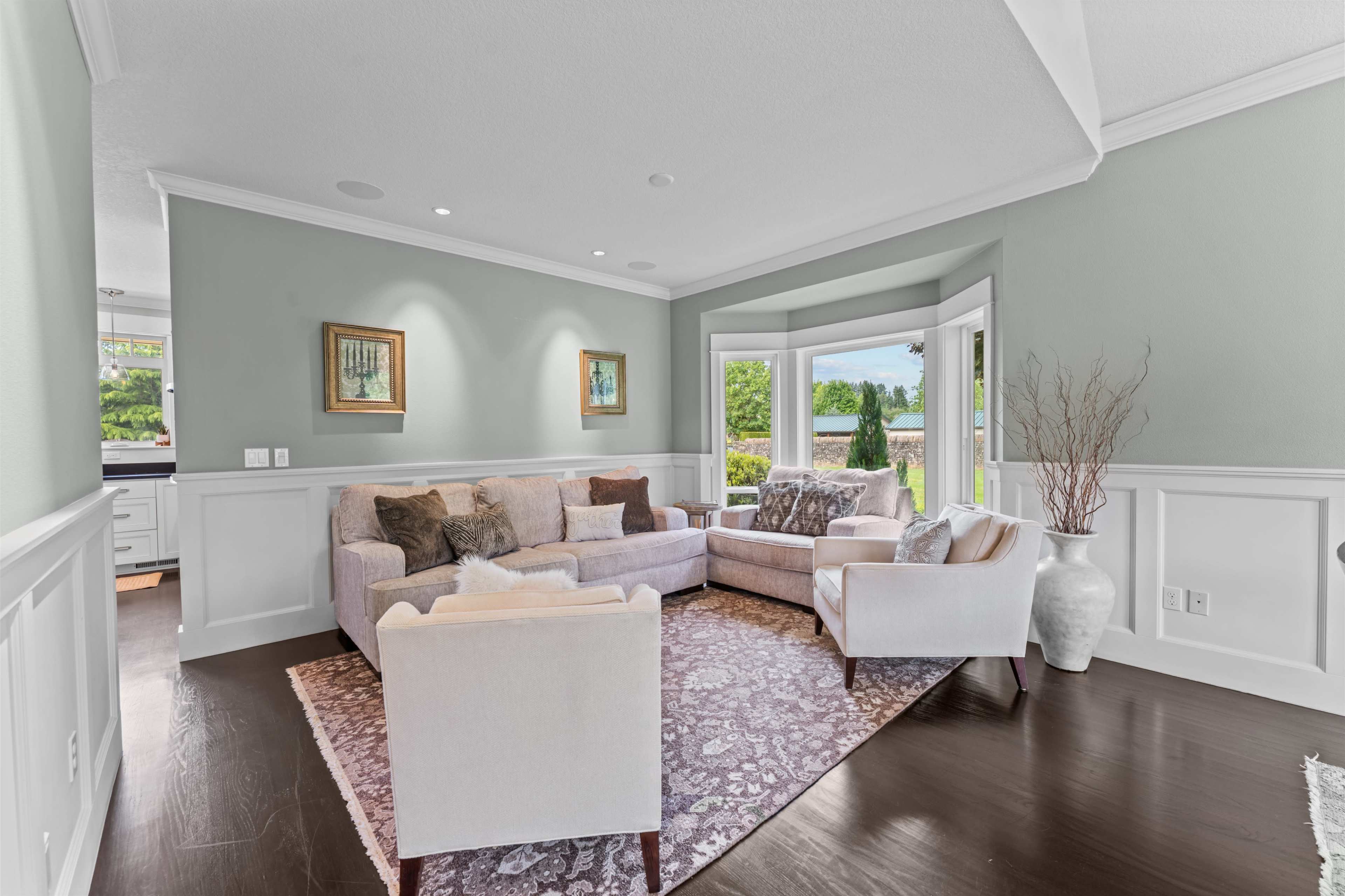 A light-colored living room features a sectional sofa, armchair, and decorative items, with large windows allowing natural light to enter.