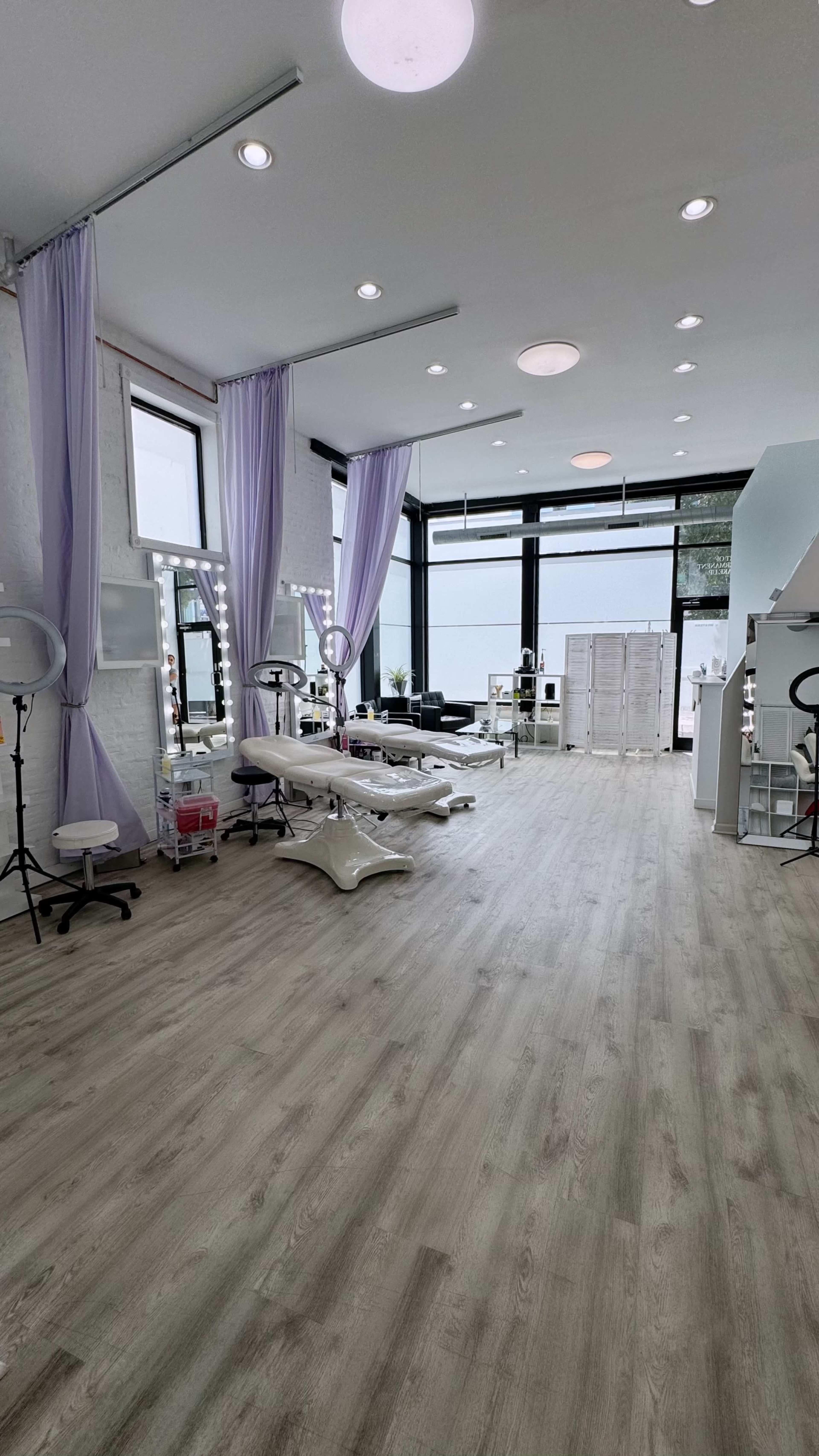 A spacious beauty studio with light-colored wooden flooring, large windows, and elegantly draped purple curtains.