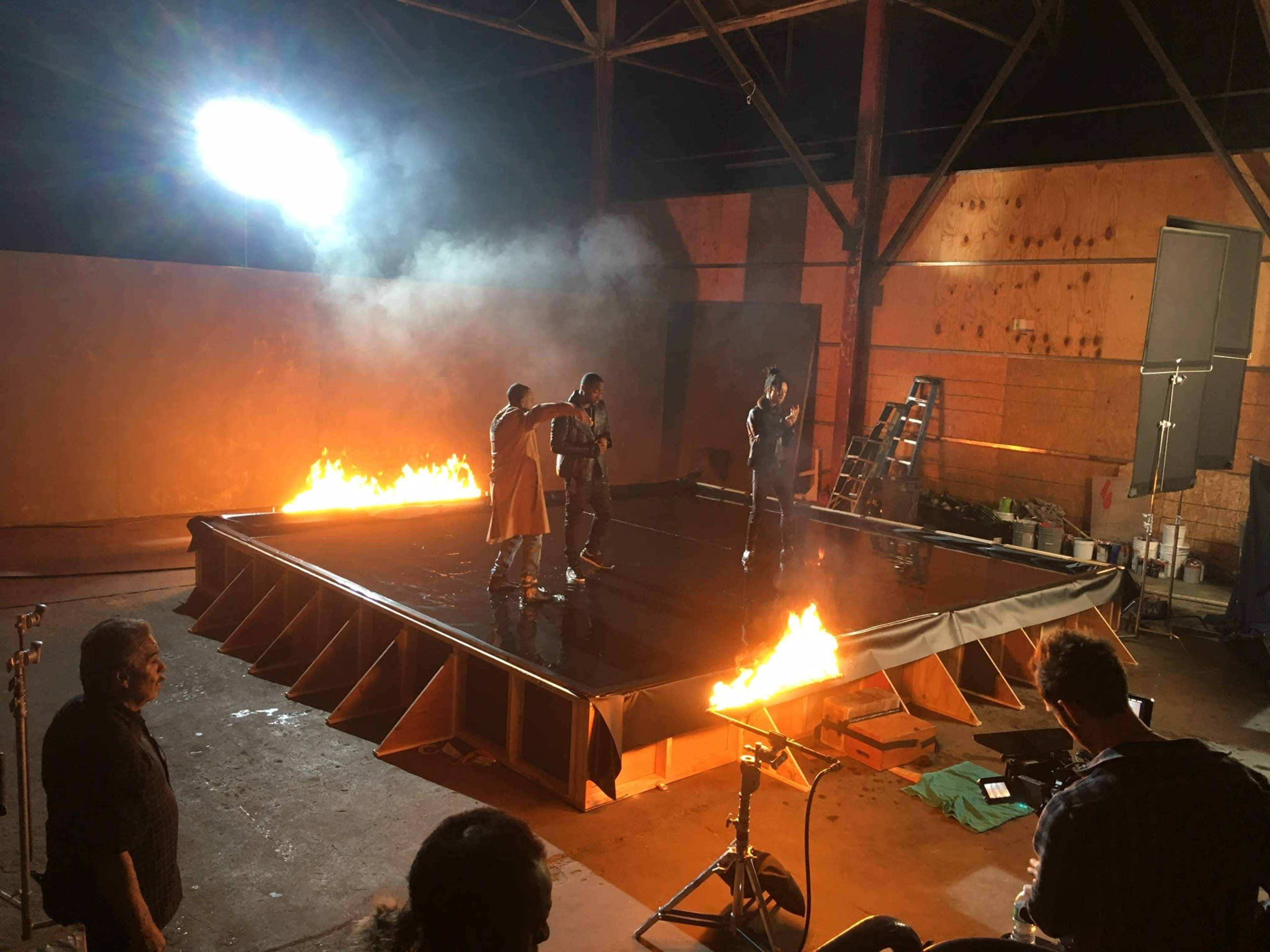 A film production set features actors performing on a platform surrounded by controlled flames, with crew members and equipment visible nearby.