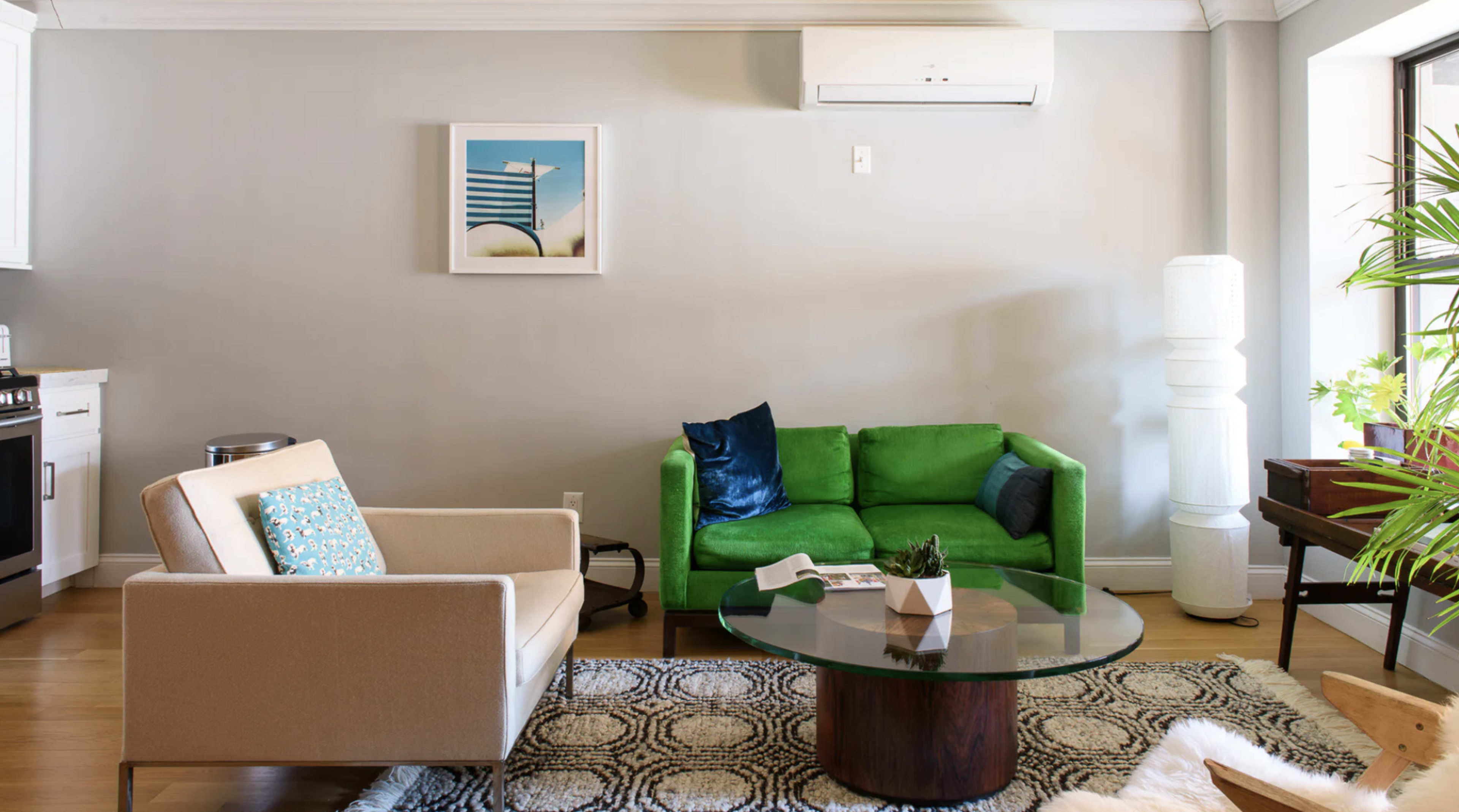 A green sofa and a beige armchair are positioned around a glass coffee table in a well-lit living room with a modern aesthetic.