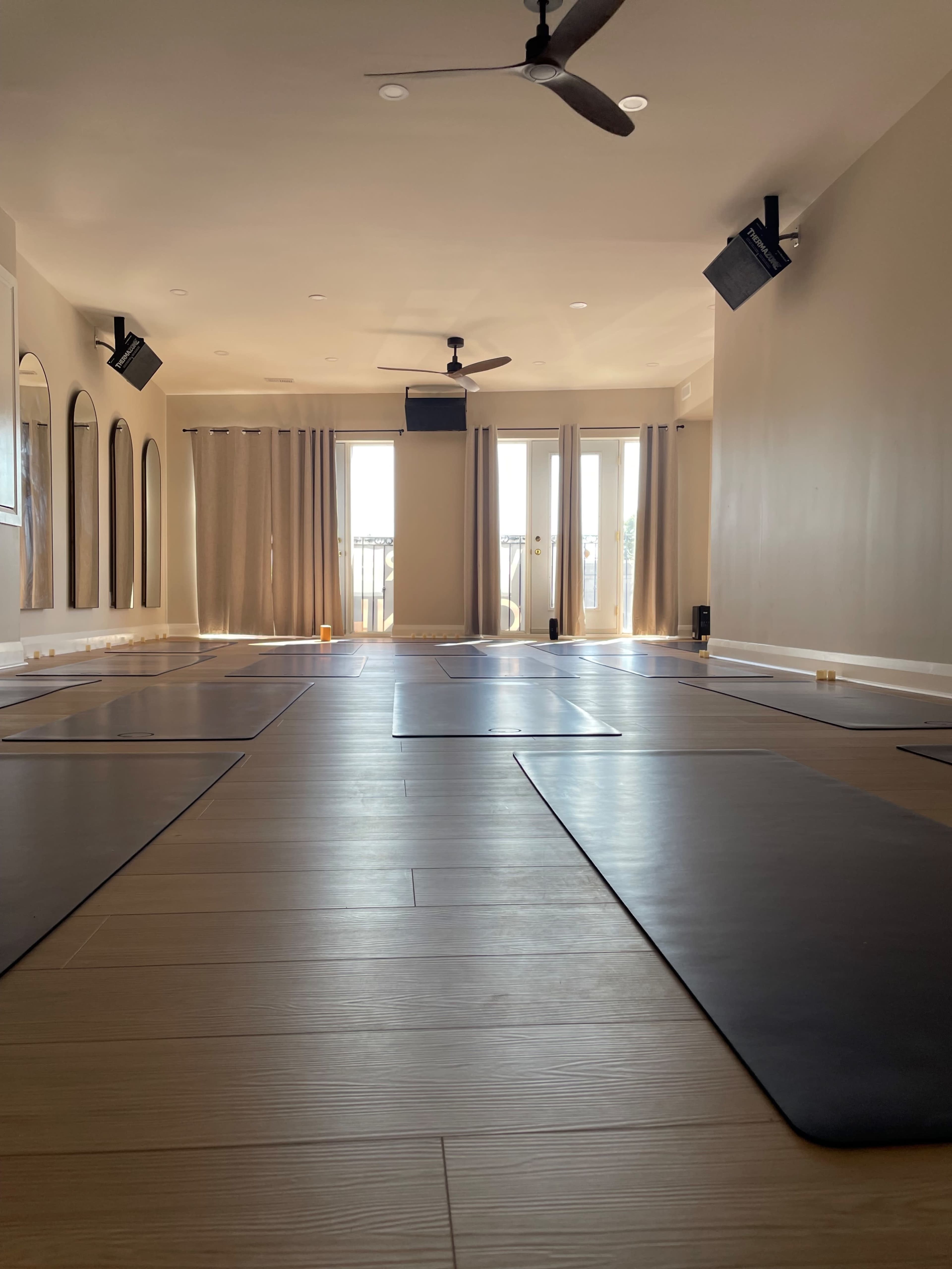 A spacious yoga studio with polished wooden floors, several yoga mats arranged on the ground, and large windows covered by light curtains.