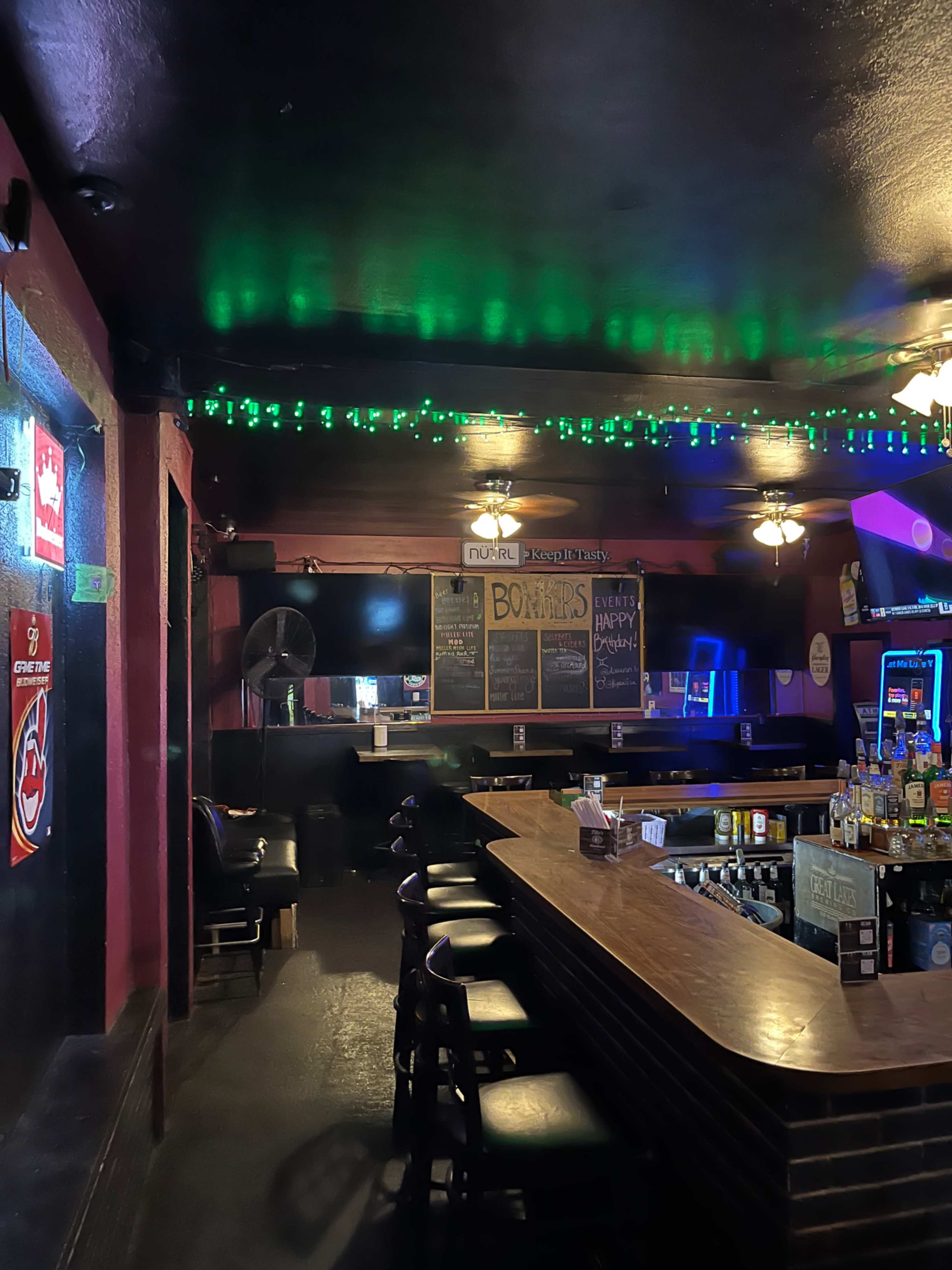 A dimly lit bar features a wooden countertop, several empty stools, and a wall adorned with menus and colorful lights.