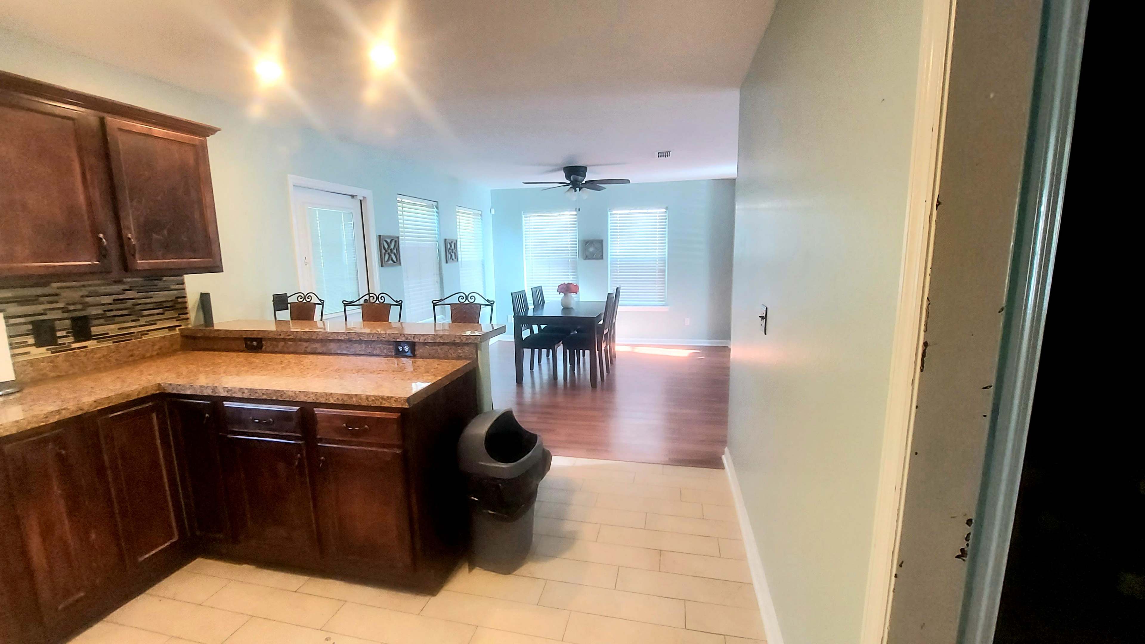 A kitchen area with brown cabinets and a granite countertop, leading into a dining space with a table and chairs under a ceiling fan.