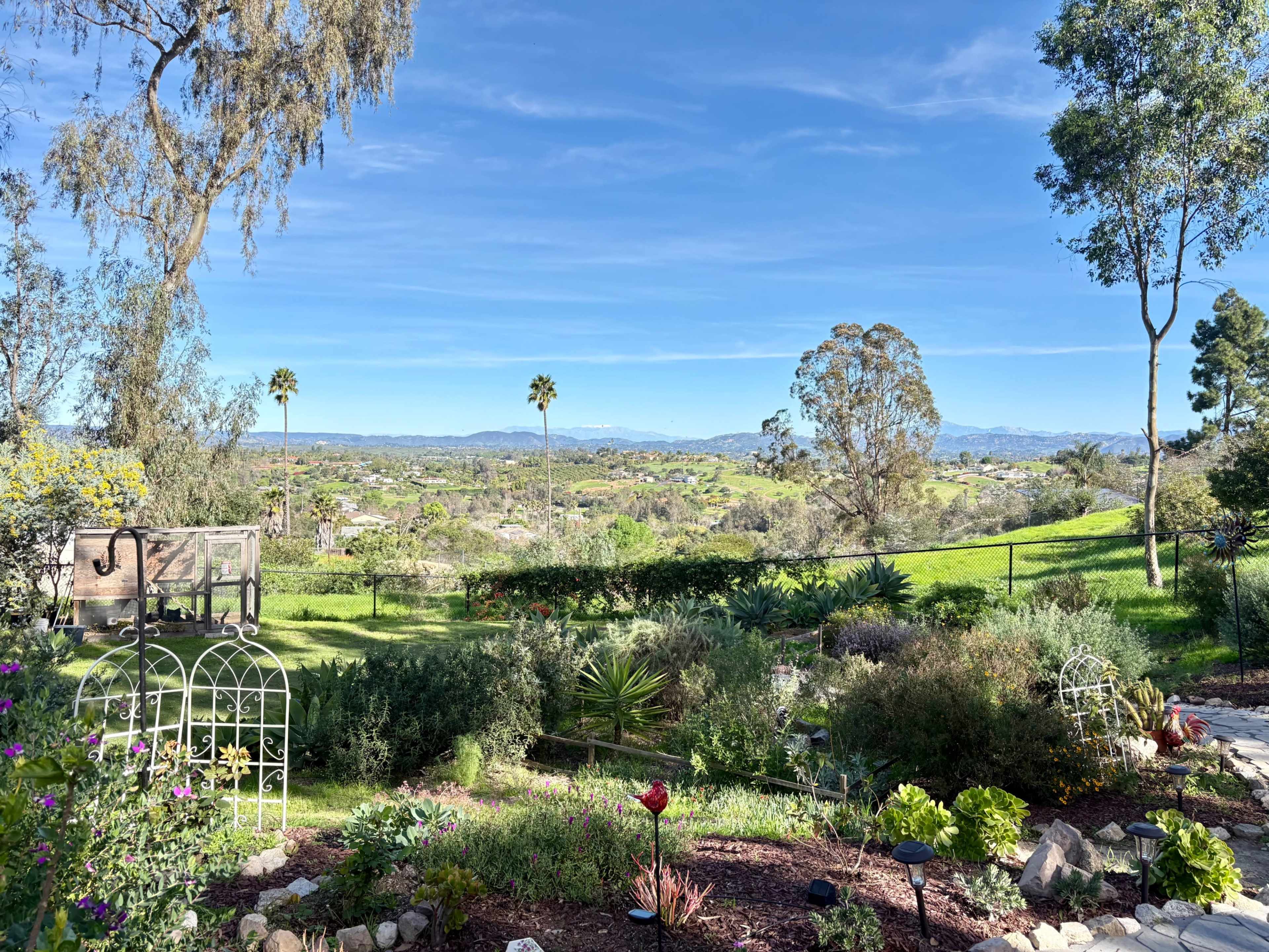 The image shows a lush garden in the foreground leading to a scenic view of rolling hills and distant mountains under a clear blue sky.