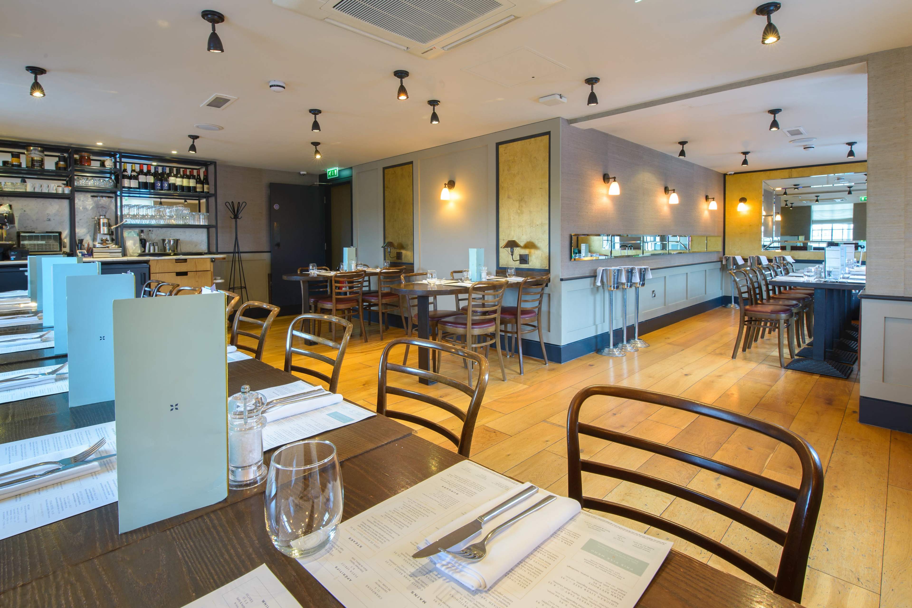 The image shows a modern restaurant interior with wooden floors, neatly arranged tables, and a bar area in the background.
