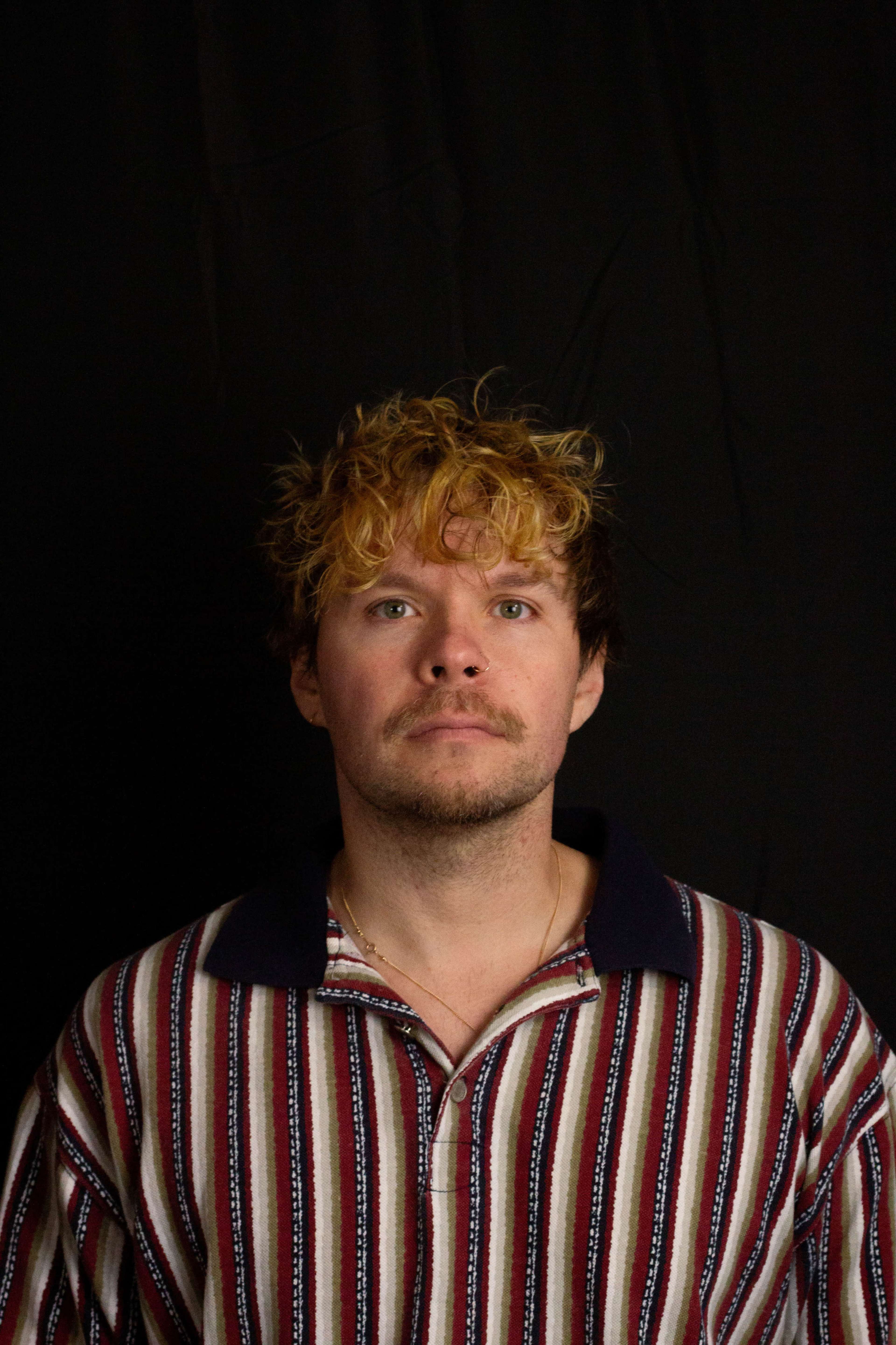 A man with curly blond hair and a striped polo shirt is standing against a dark background.