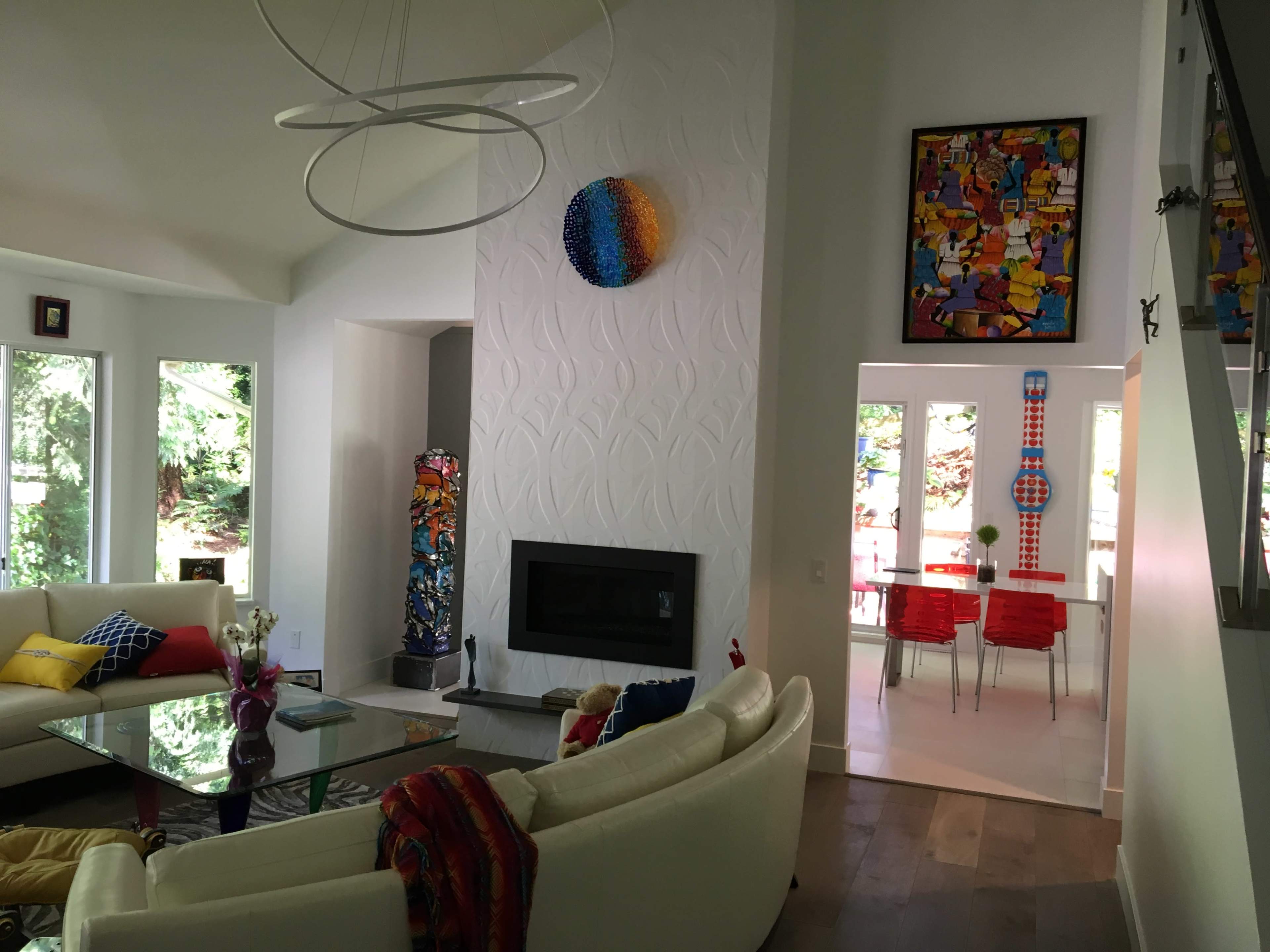 A bright, modern living room with a circular white sofa, a glass coffee table, a fireplace, and colorful artwork on the walls, leading into a dining area with red chairs.