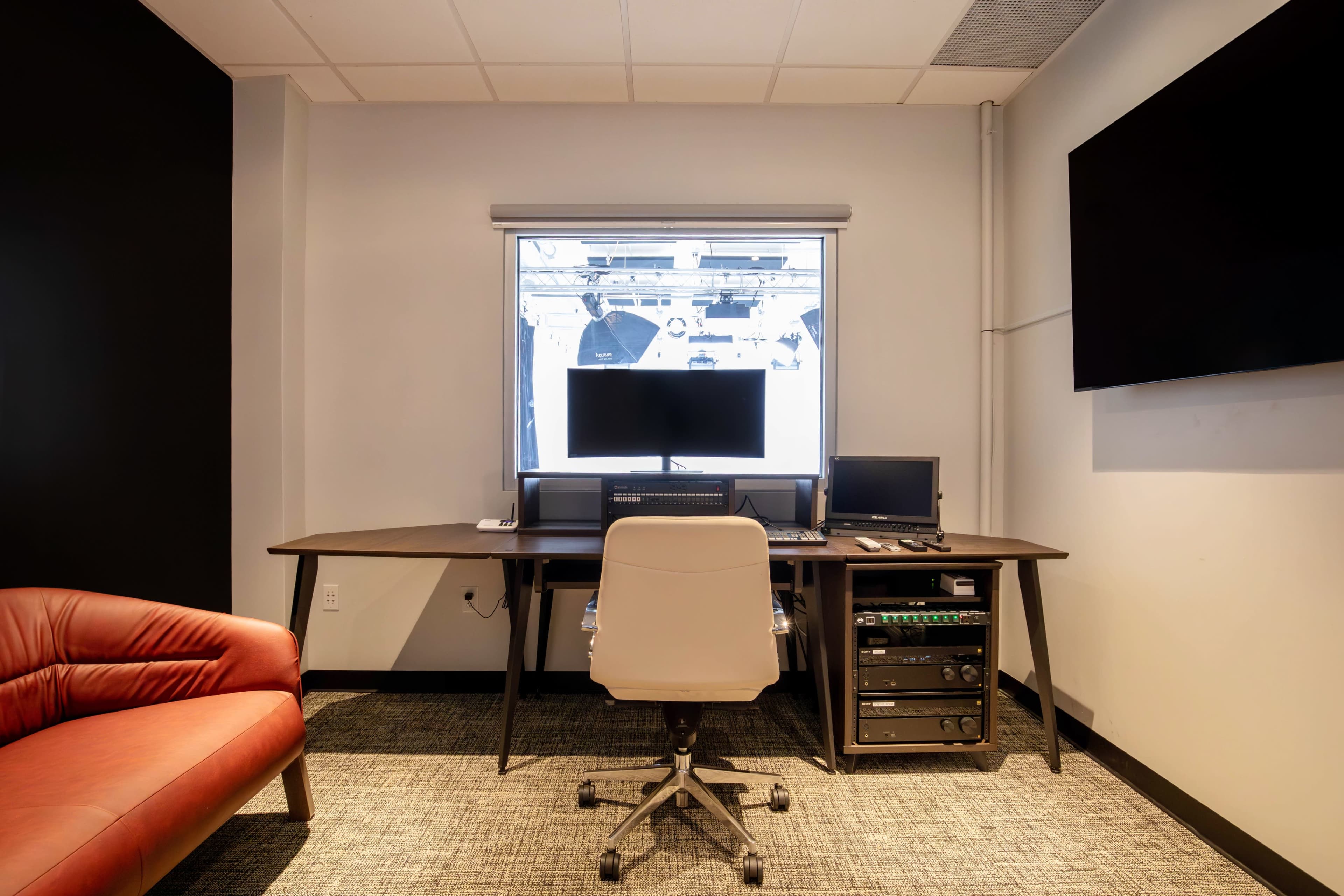 The image shows a modern office workspace featuring a desk with multiple computer monitors, an ergonomic chair, and a red lounge chair against a black wall.