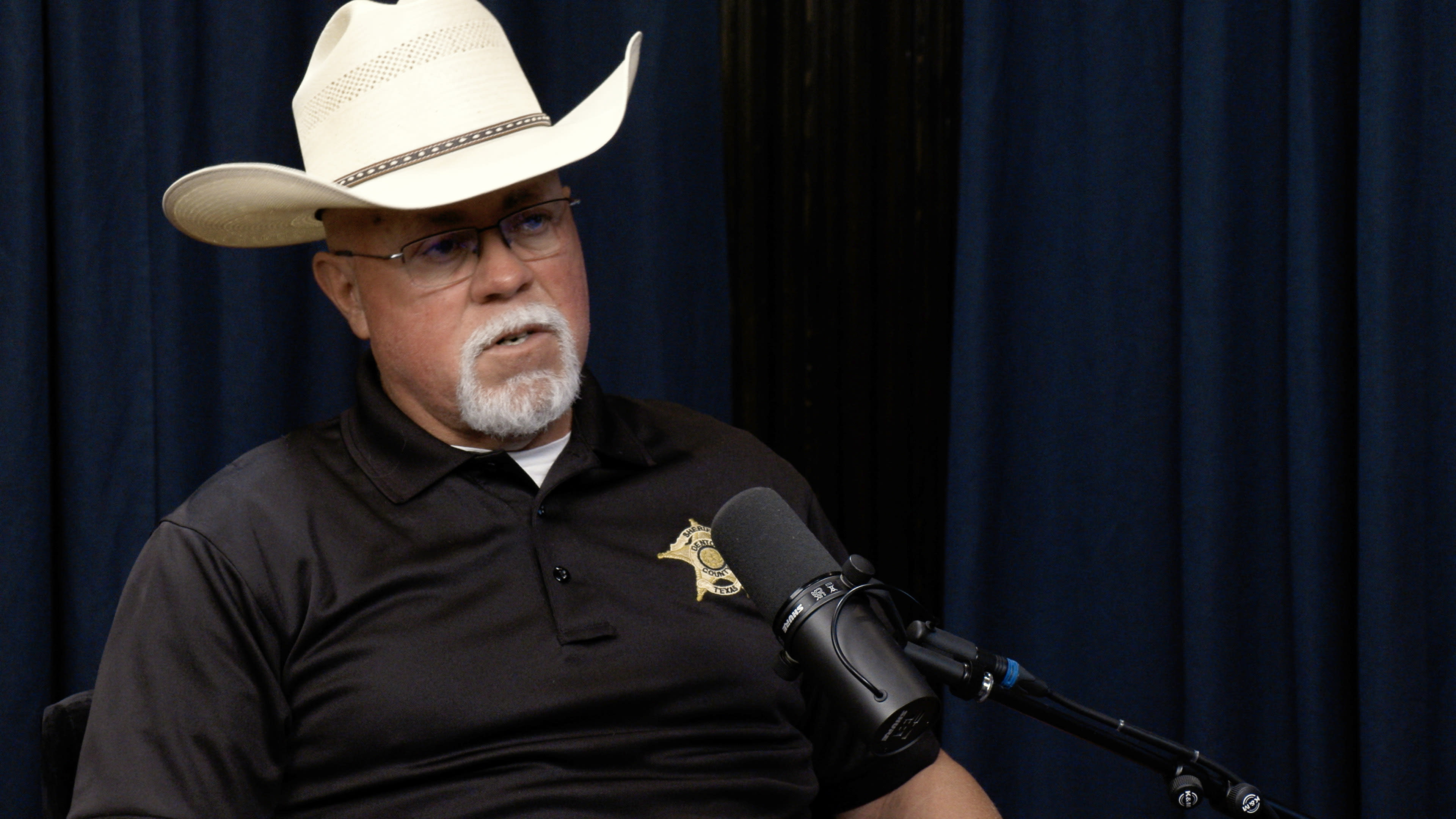 A man in a cowboy hat and a black polo shirt with a badge sits in front of a microphone against a blue backdrop.