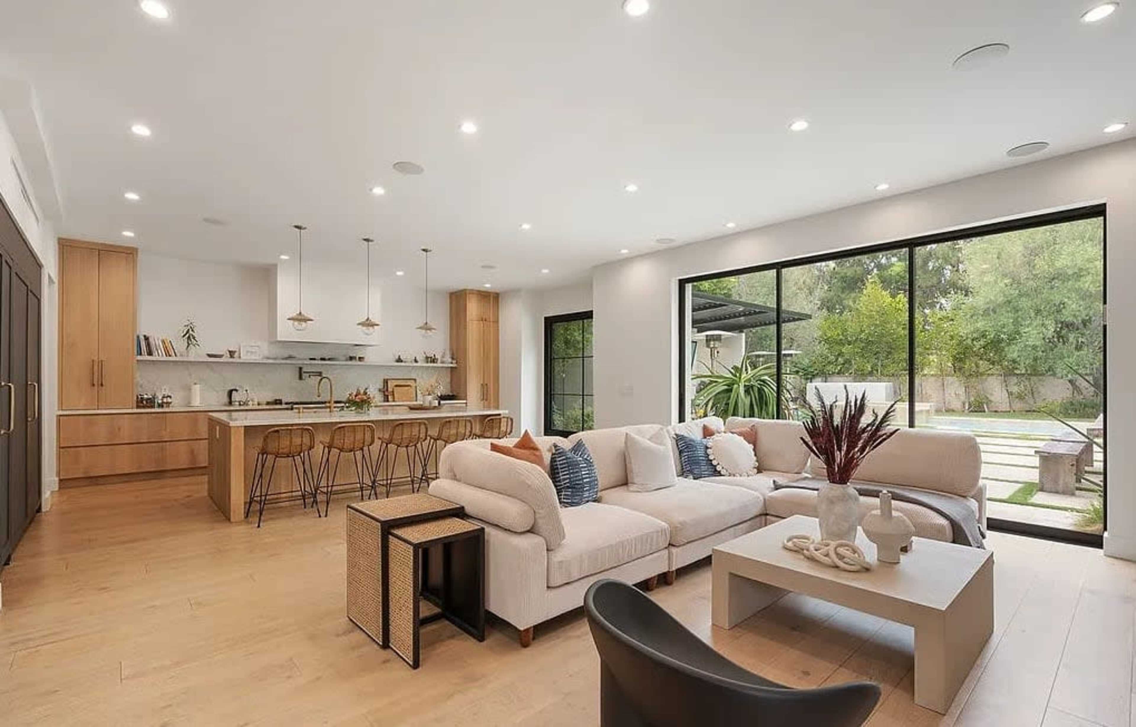 The image shows a modern living room and kitchen area with light wood flooring, a large sectional sofa, and a spacious window overlooking a green garden.