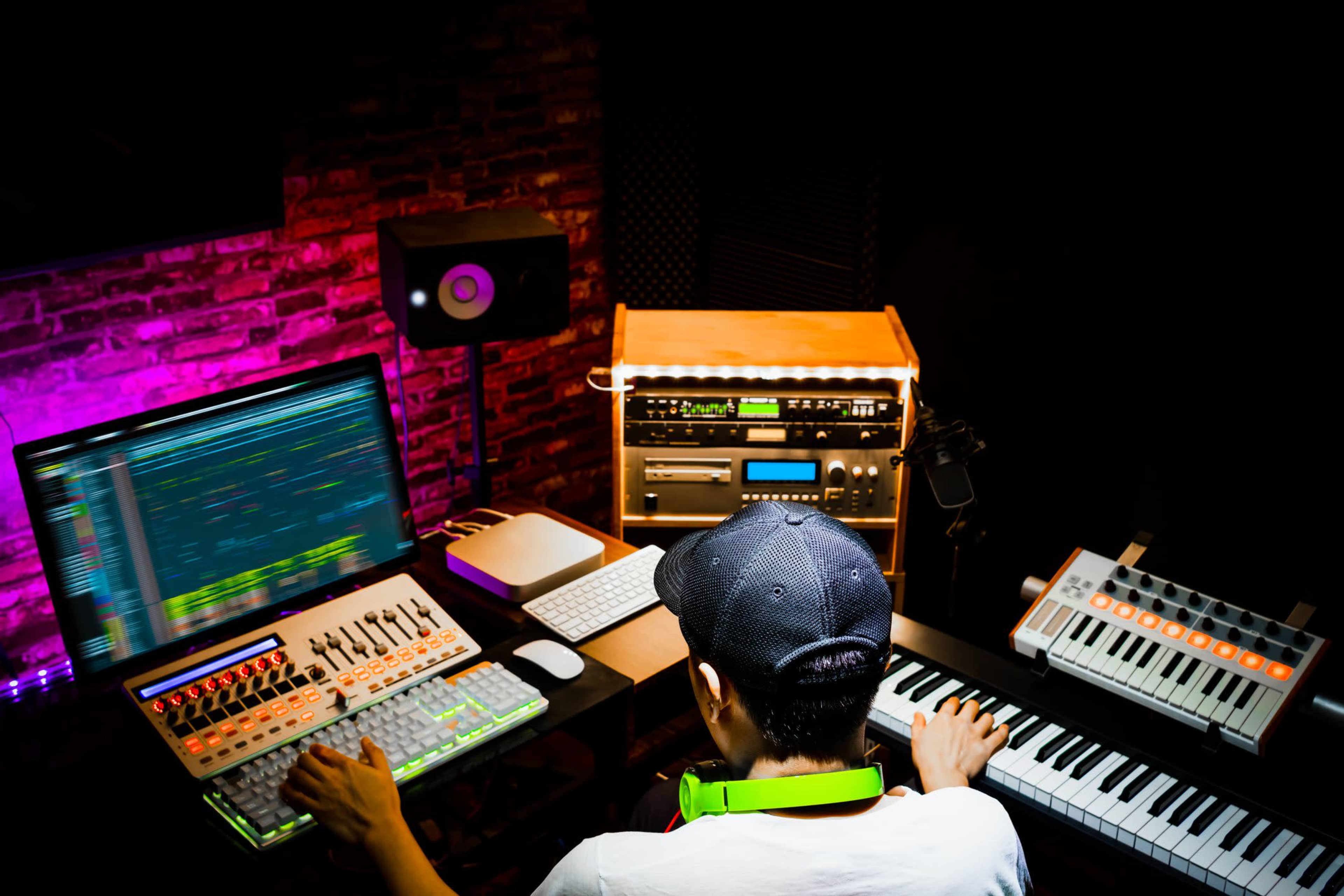 A person is seated at a desk with a computer, producing music using a digital audio workstation, synthesizers, and a MIDI keyboard in a dimly lit studio.