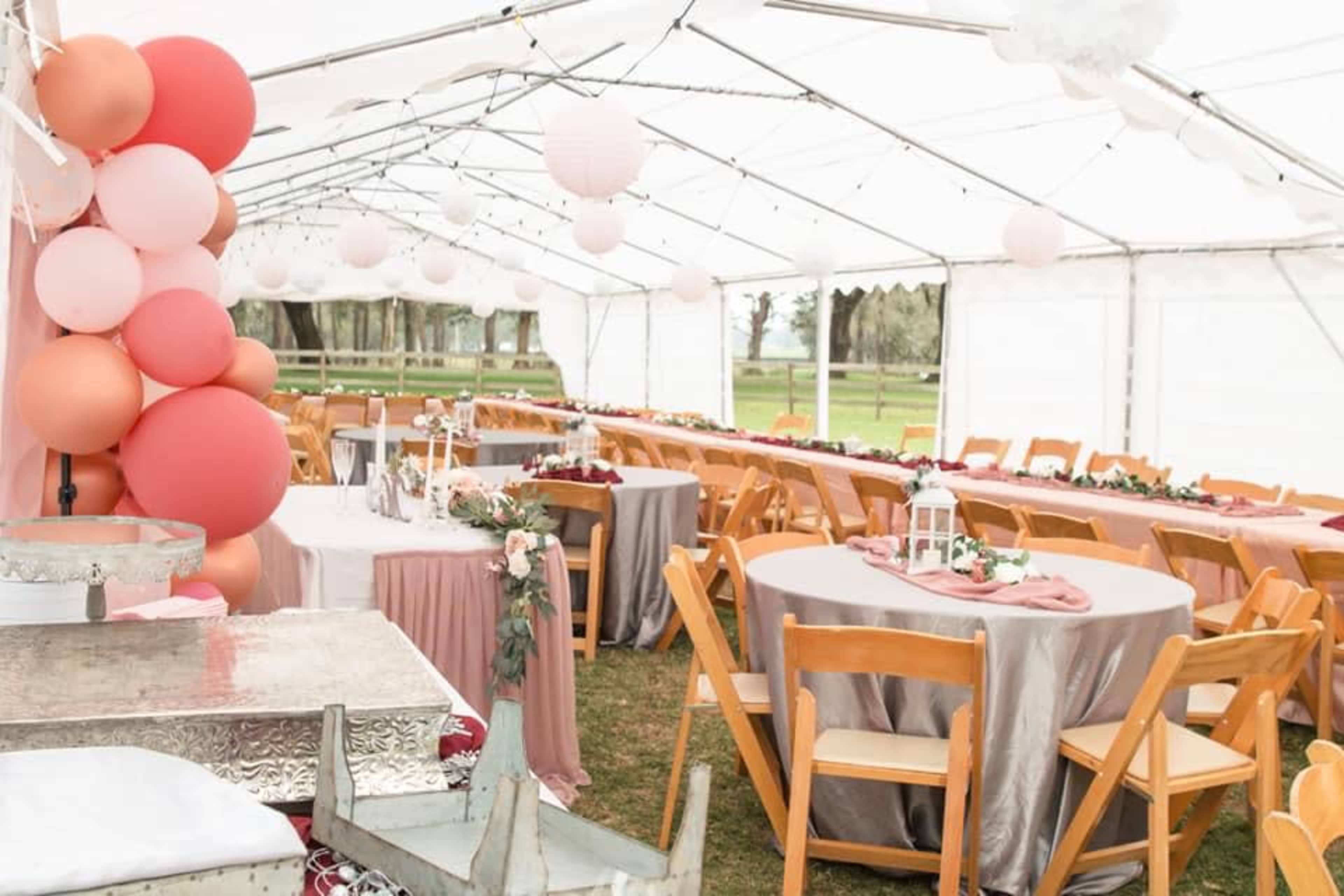 A large white tent is set up with round and rectangular tables, decorated with linens and centerpieces, alongside a display of pink and peach balloons.