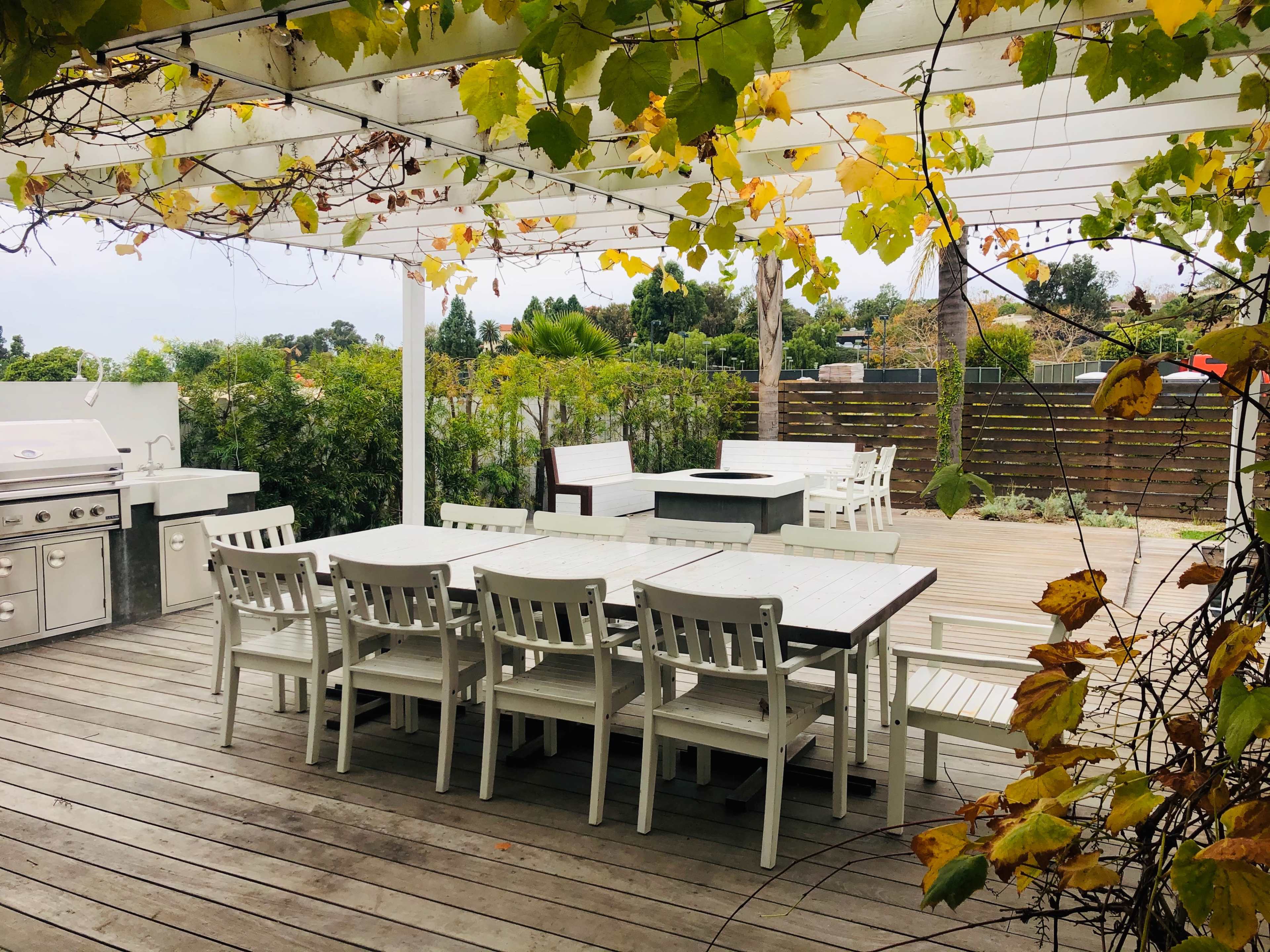 A spacious outdoor dining area features white wooden tables and chairs under a pergola, surrounded by greenery and vines.